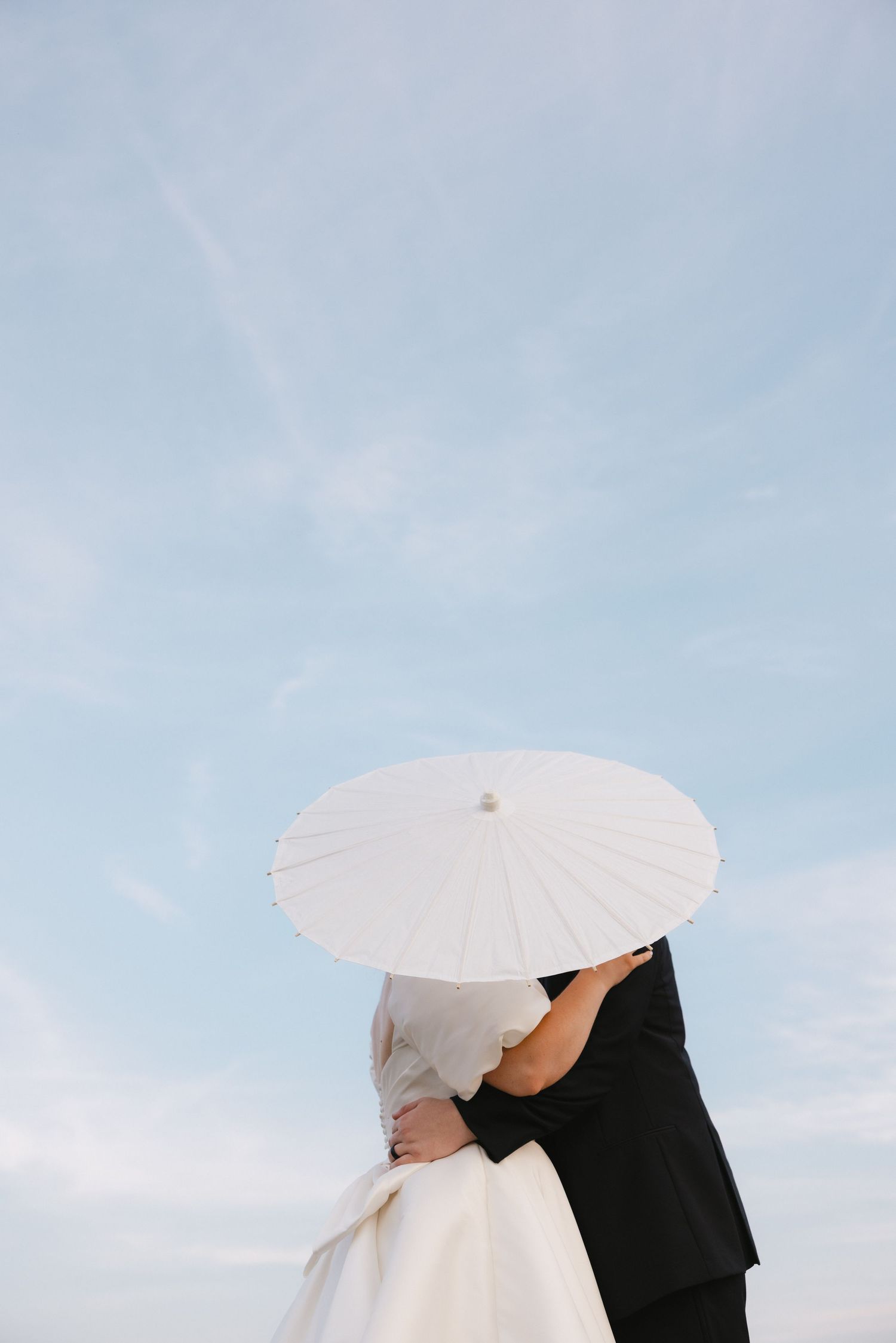 A romantic silhouette of newlyweds sharing a moment under a white parasol against blue sky.