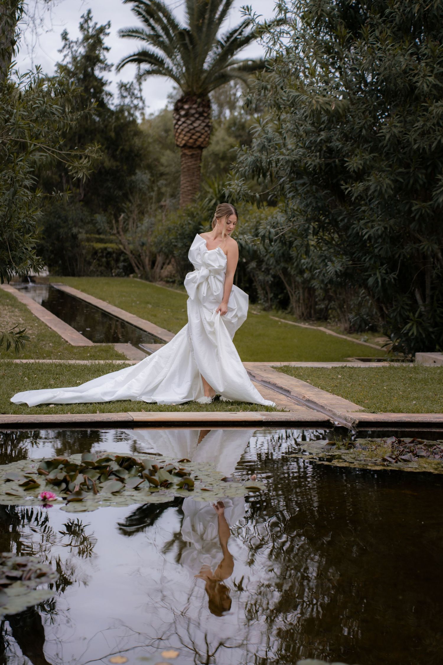 A bride in a flowing white gown poses reflectively by a scenic pond with water lilies.