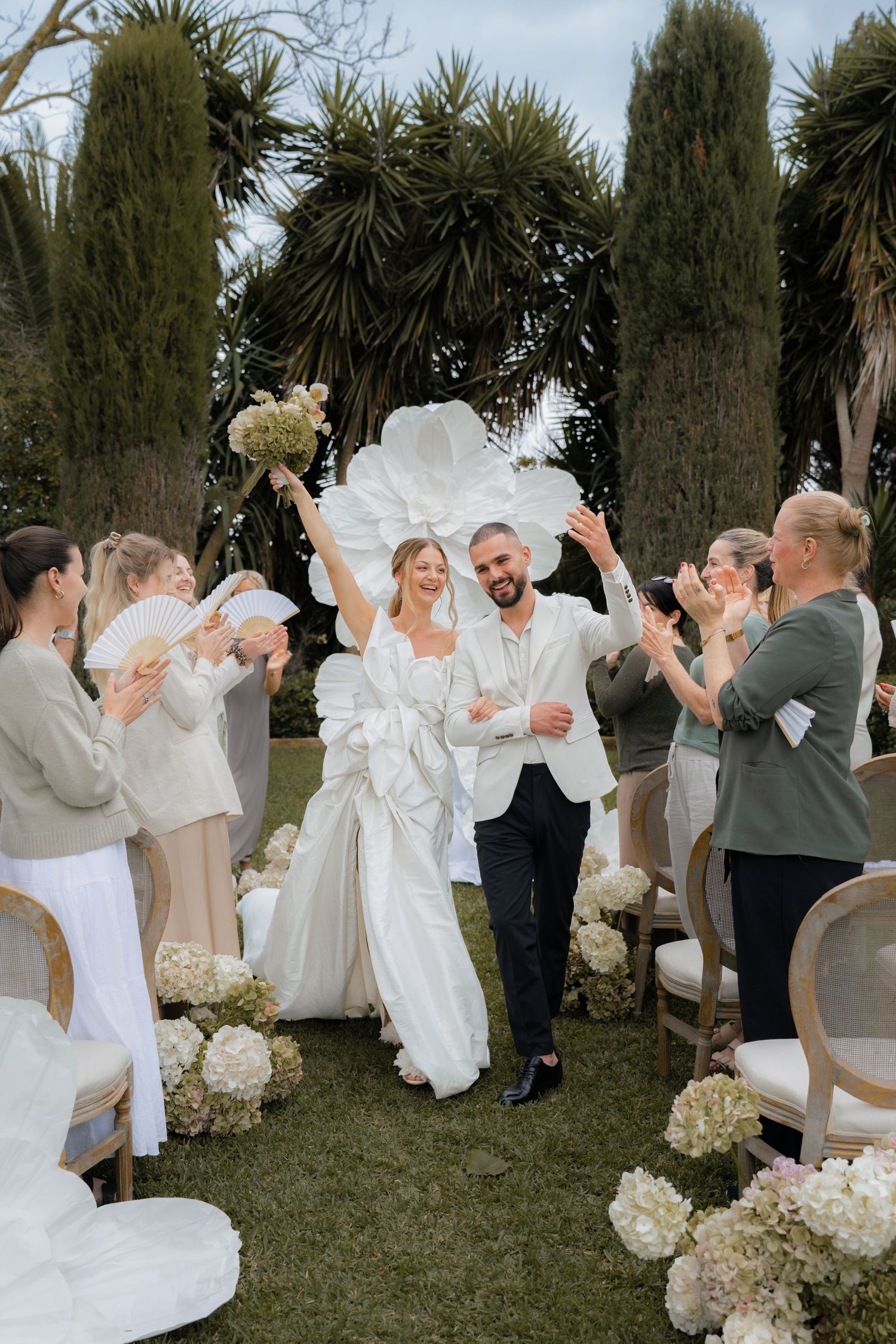 Wedding party in all-white attire celebrating outdoors among tropical plants and flowers.