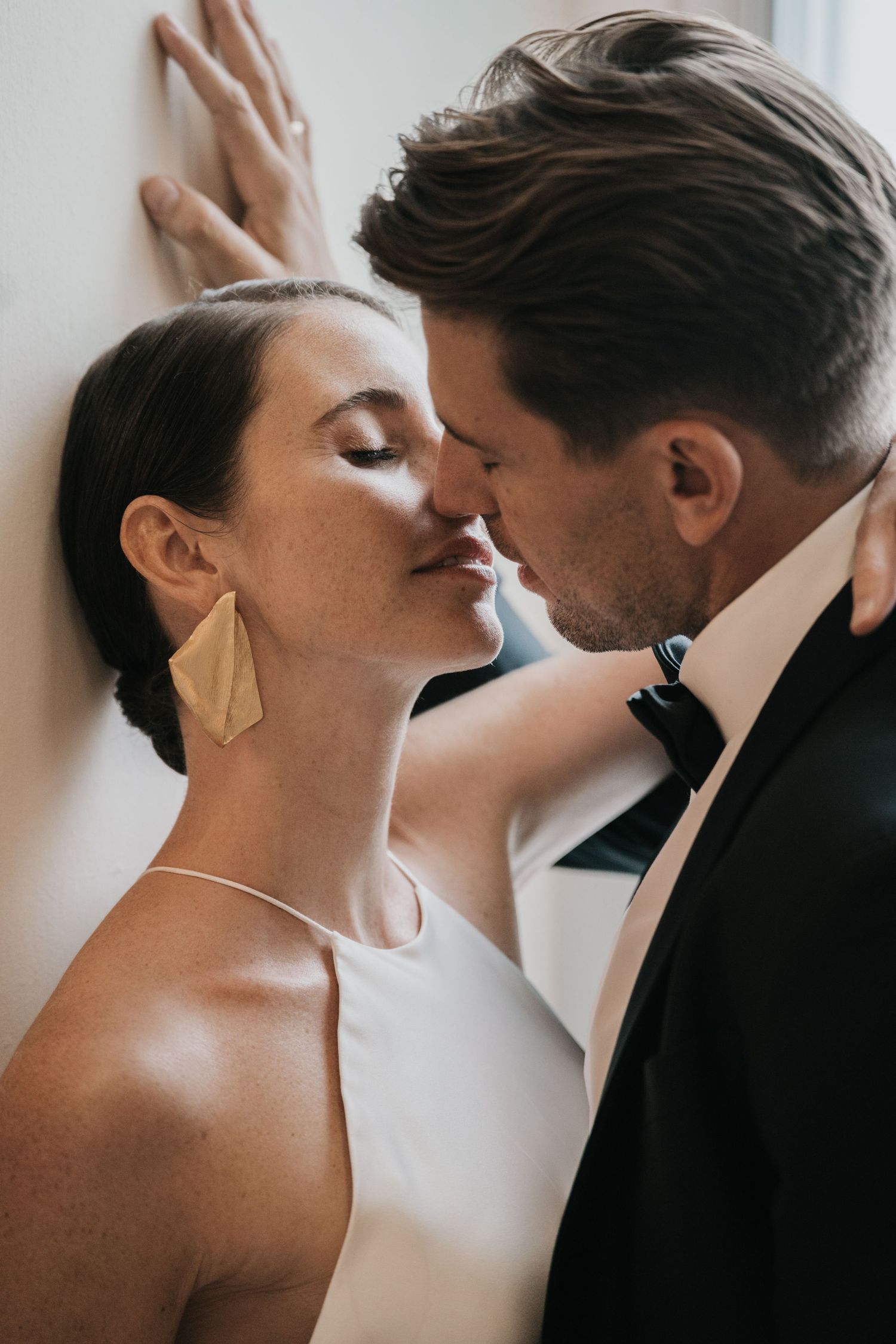 An intimate moment between a couple in formal attire against a white wall.