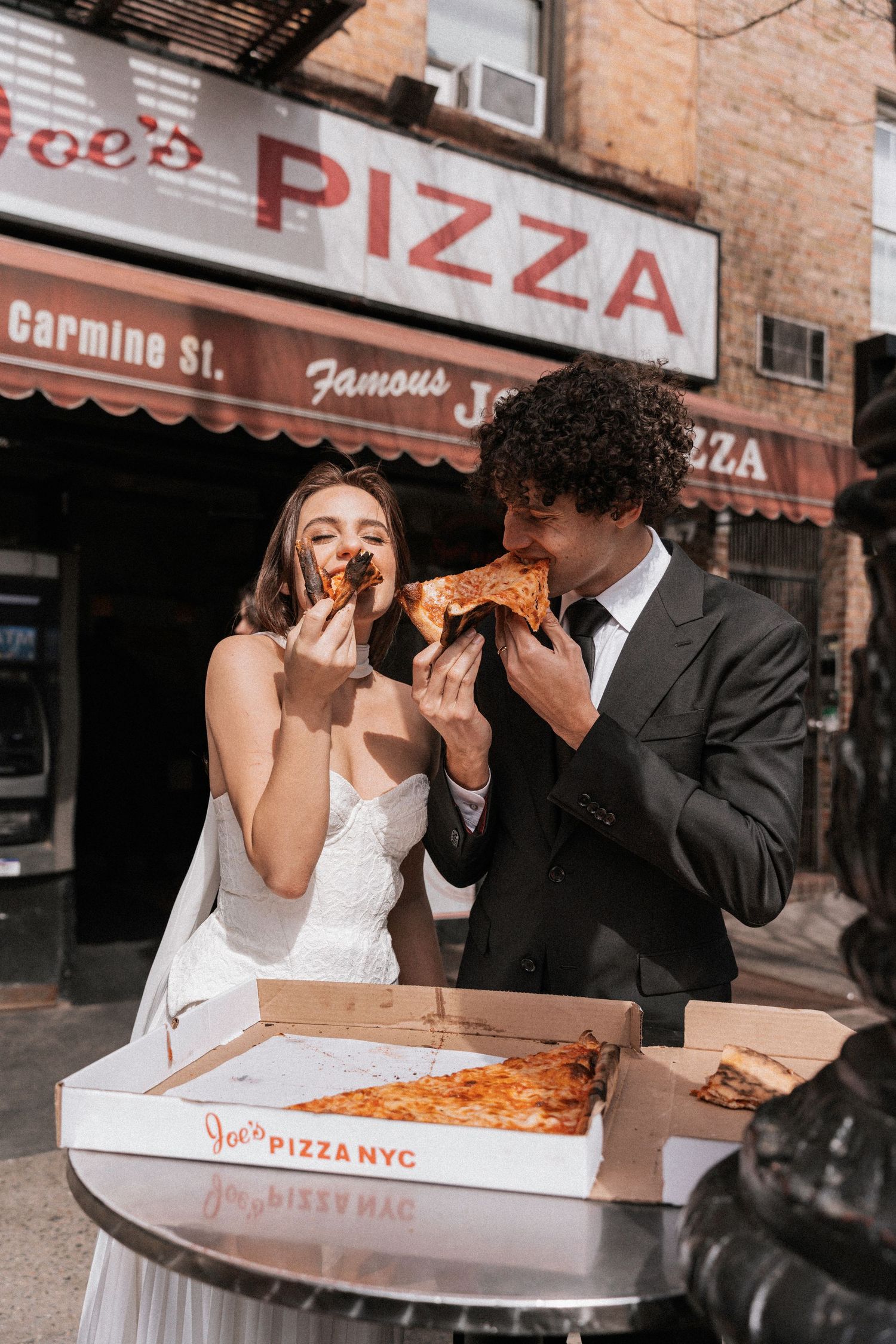 A couple in wedding attire shares slices of New York pizza outside a pizzeria.