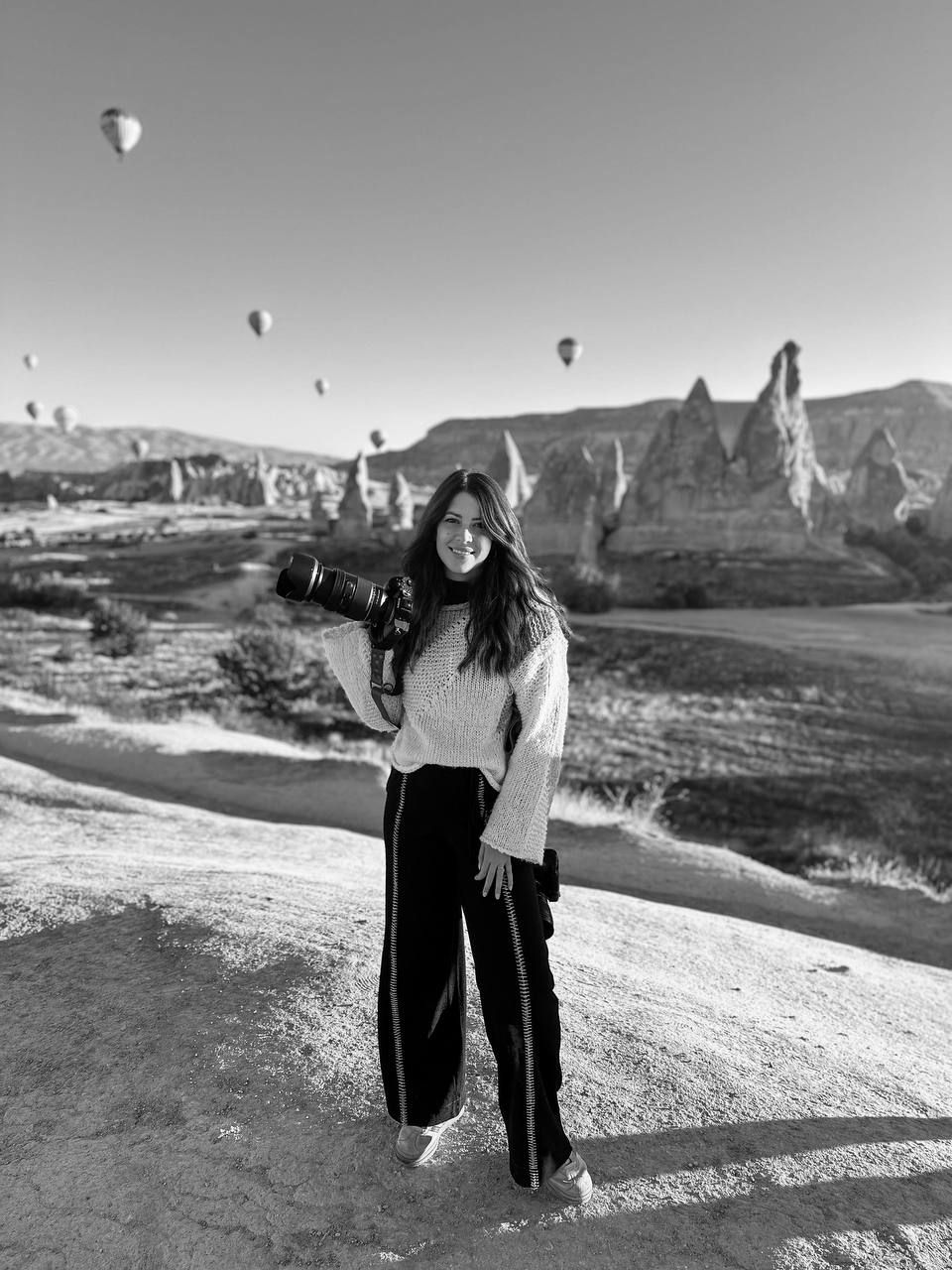 Hot air balloons float above desert landscape while people admire scenic mountain views from a rocky overlook at sunset.