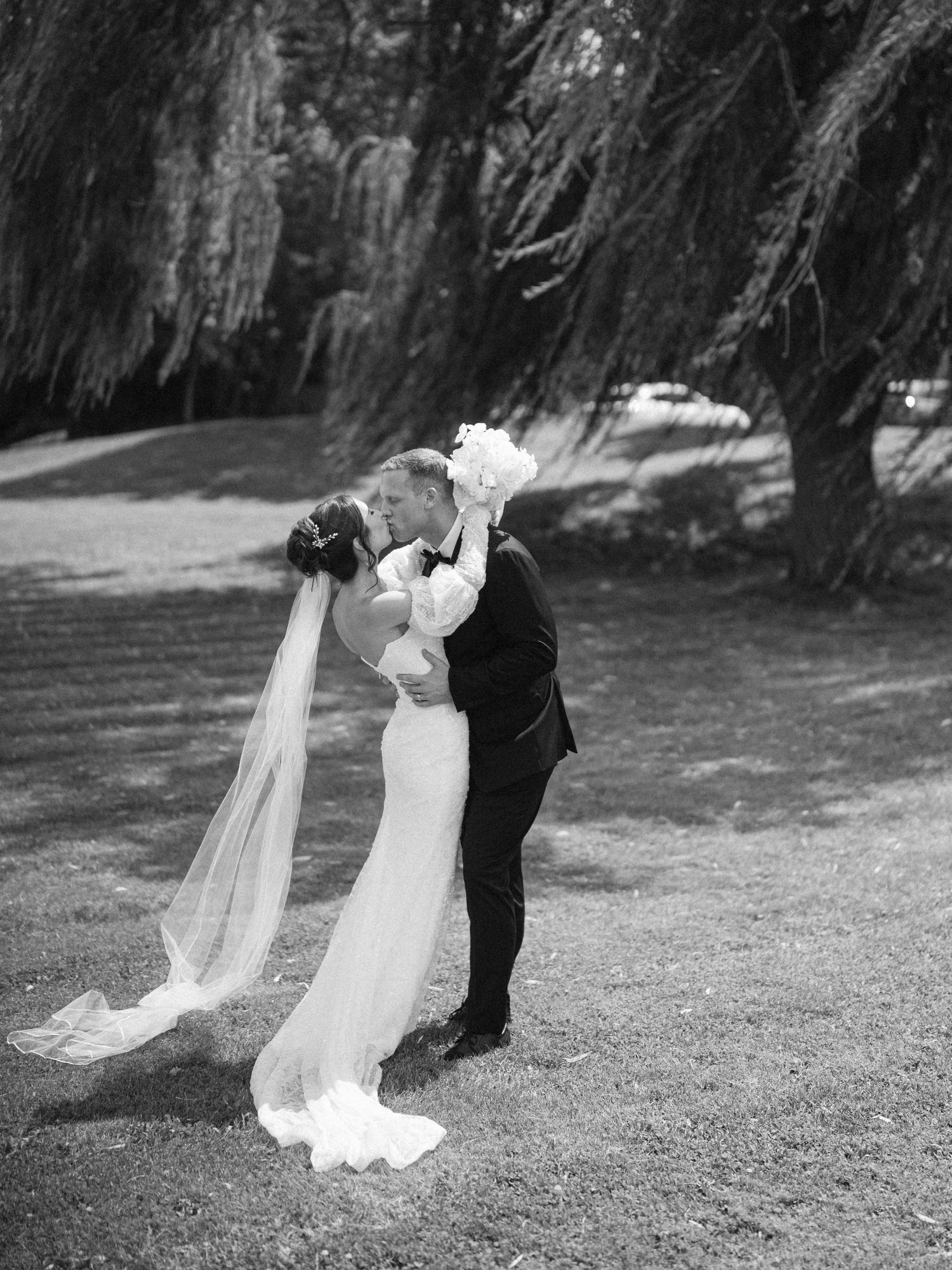 Black and white photo of wedding couple in dramatic dip pose under willow trees.