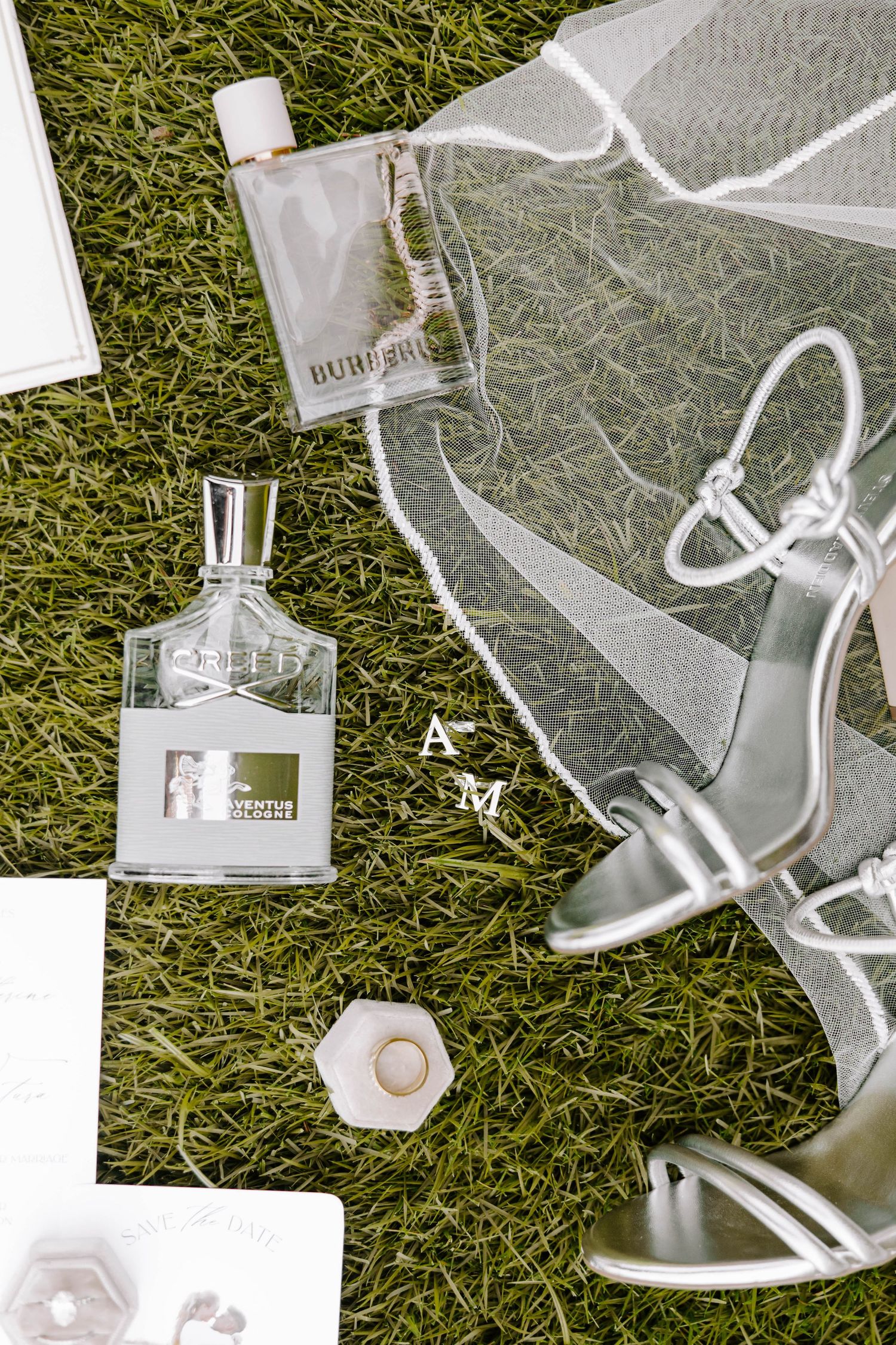 Wedding flat lay on green grass with Burberry and Creed perfume bottles, silver strappy heels, a sheer veil, a ring box and AM monogram.