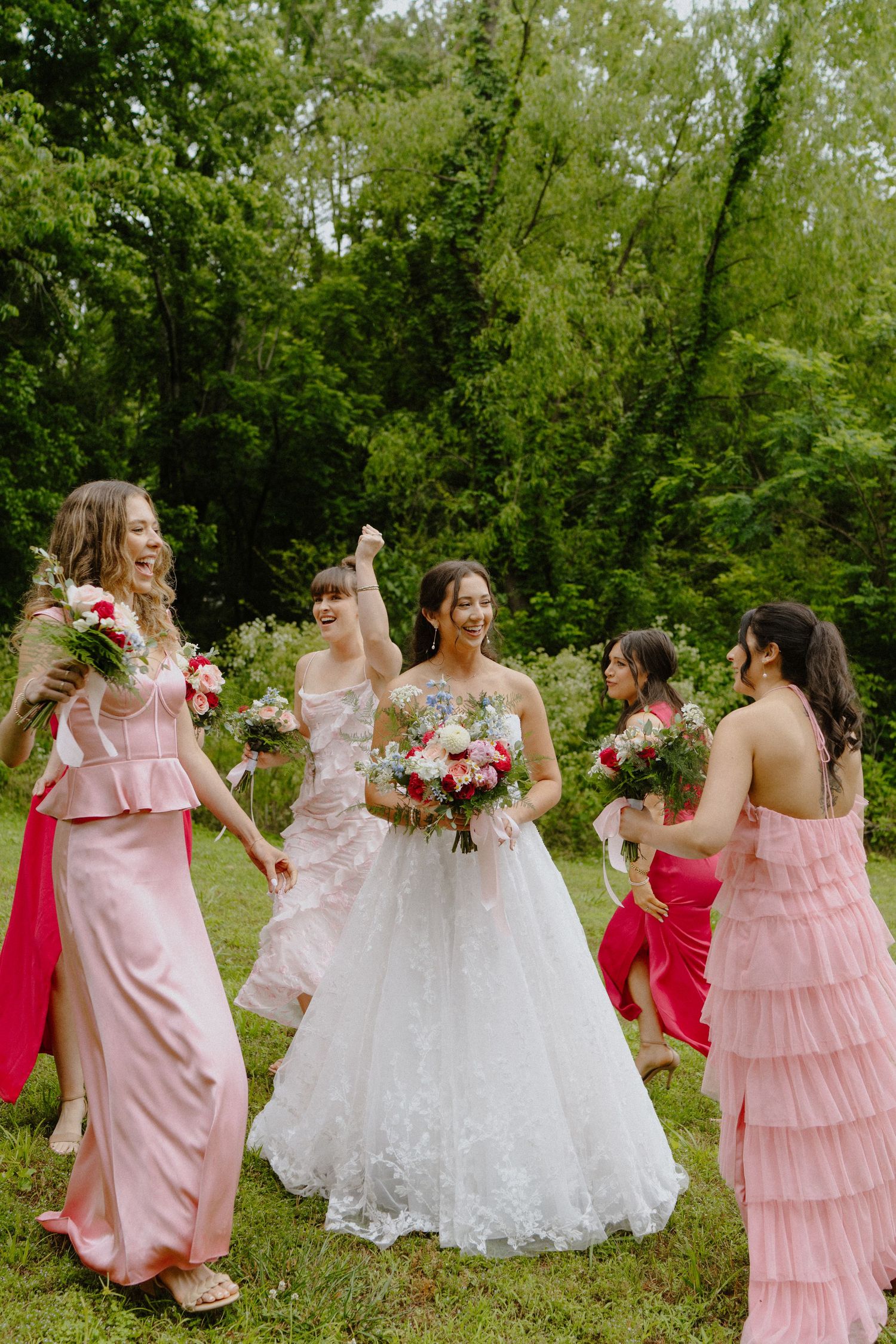 Bridesmaids in pink dresses dance and celebrate together on a sunny outdoor lawn.