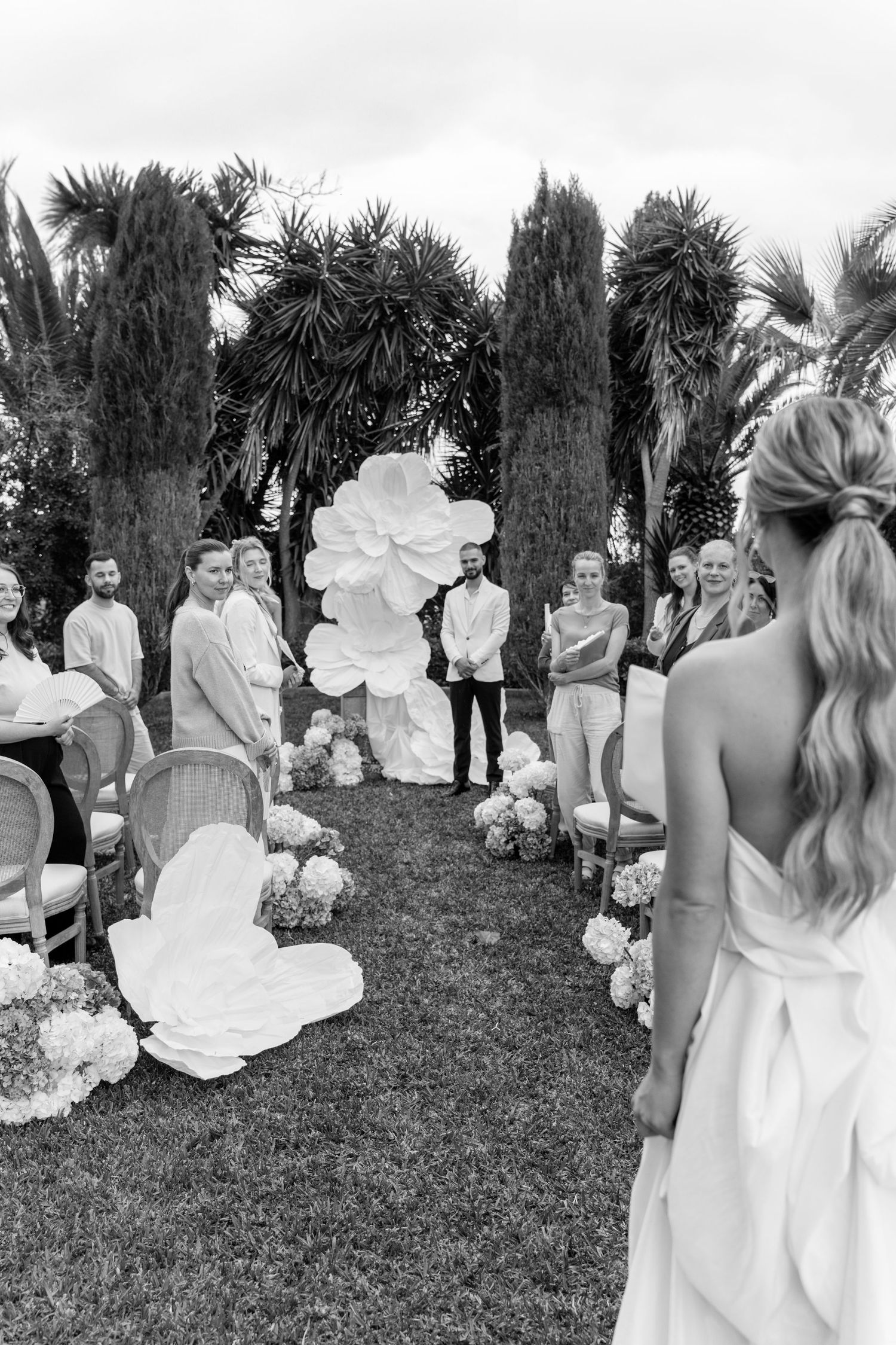 Black and white photo of wedding ceremony guests gathered outdoors among cypress trees.