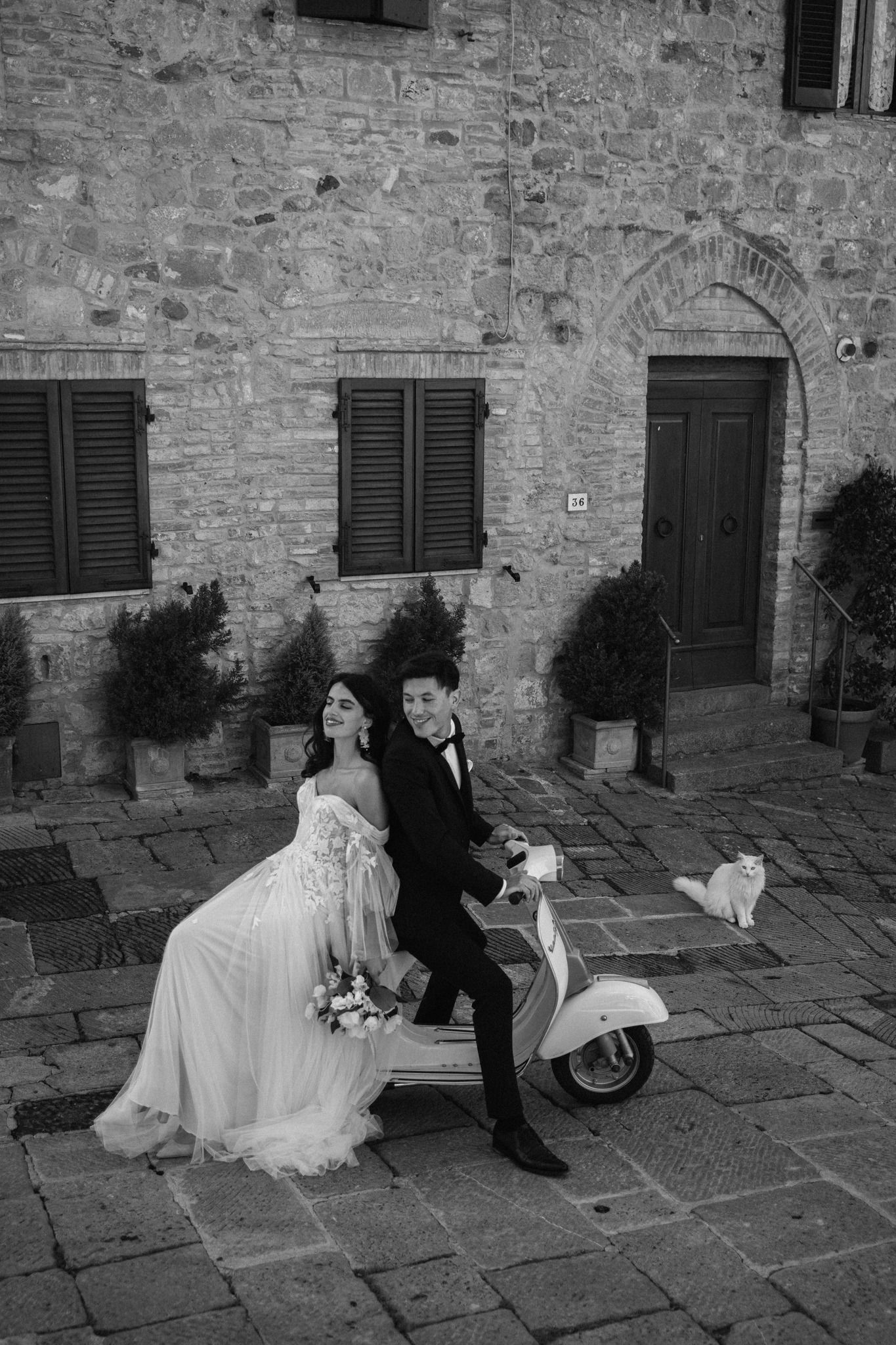 A stylish wedding couple poses with a vintage Vespa scooter against an Italian stone building.
