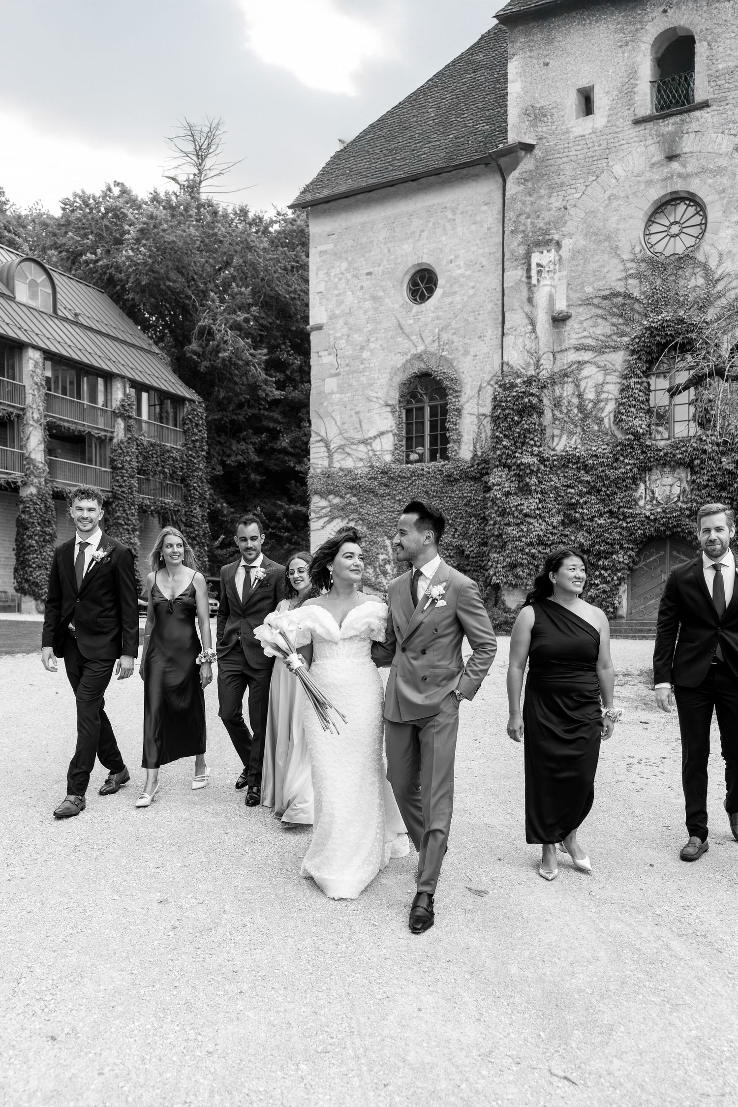 A wedding party walks in a line formation outside a historic stone building.