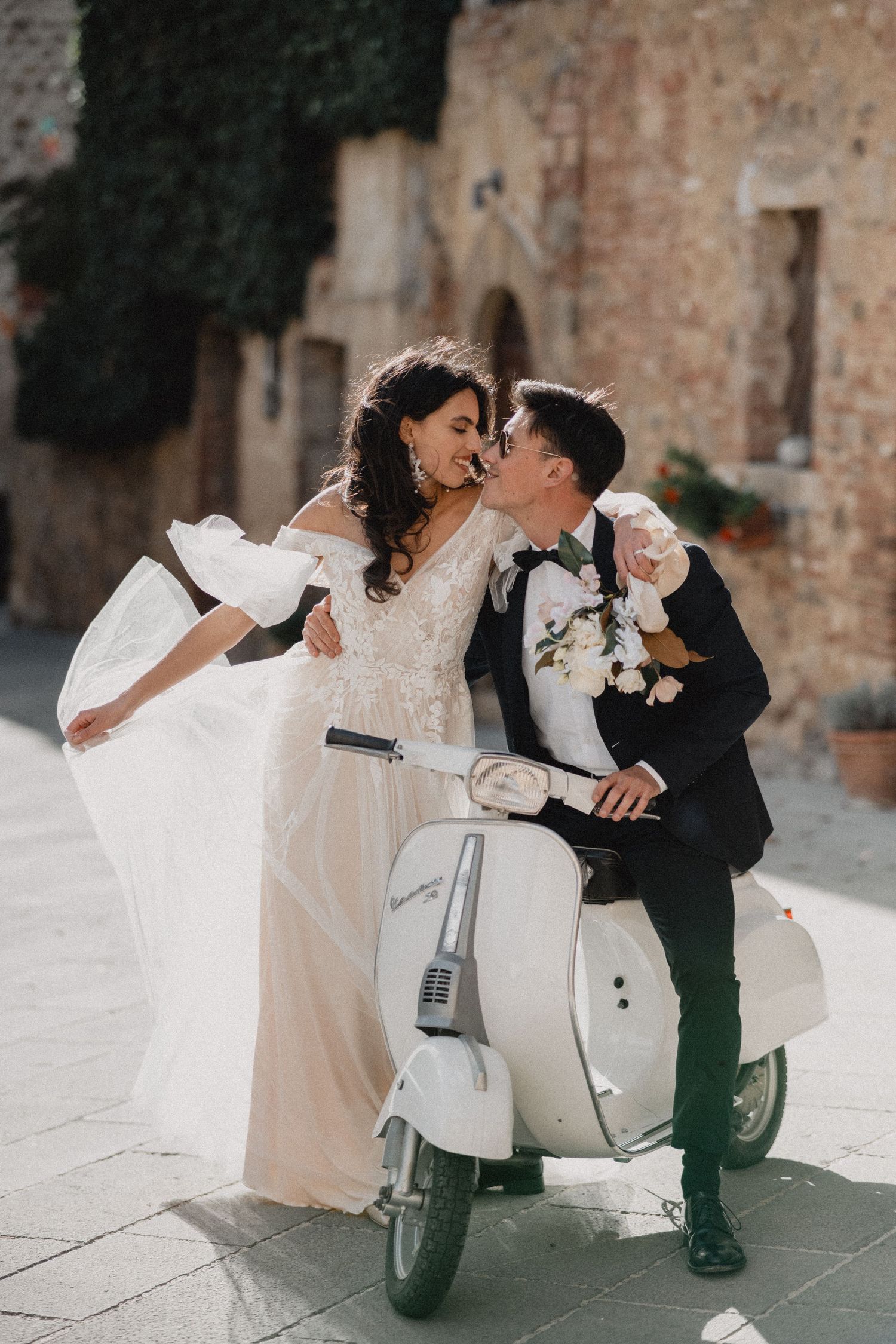 A playful wedding couple shares a romantic moment on a vintage Vespa in an Italian courtyard.