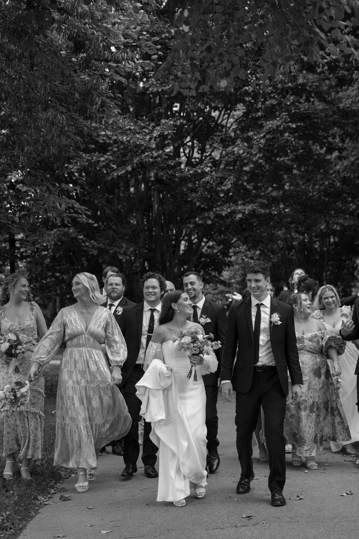 Wedding party walking and laughing together on a tree-lined path.
