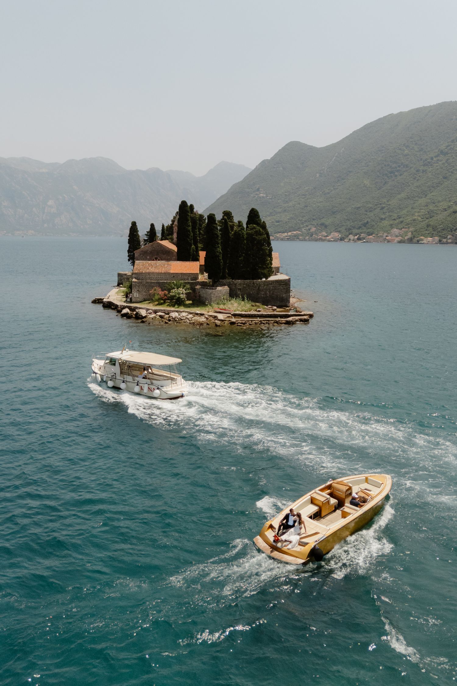 Two motorboats cruise through turquoise bay waters past a small island with a stone church and cypress trees, framed by misty mountains.