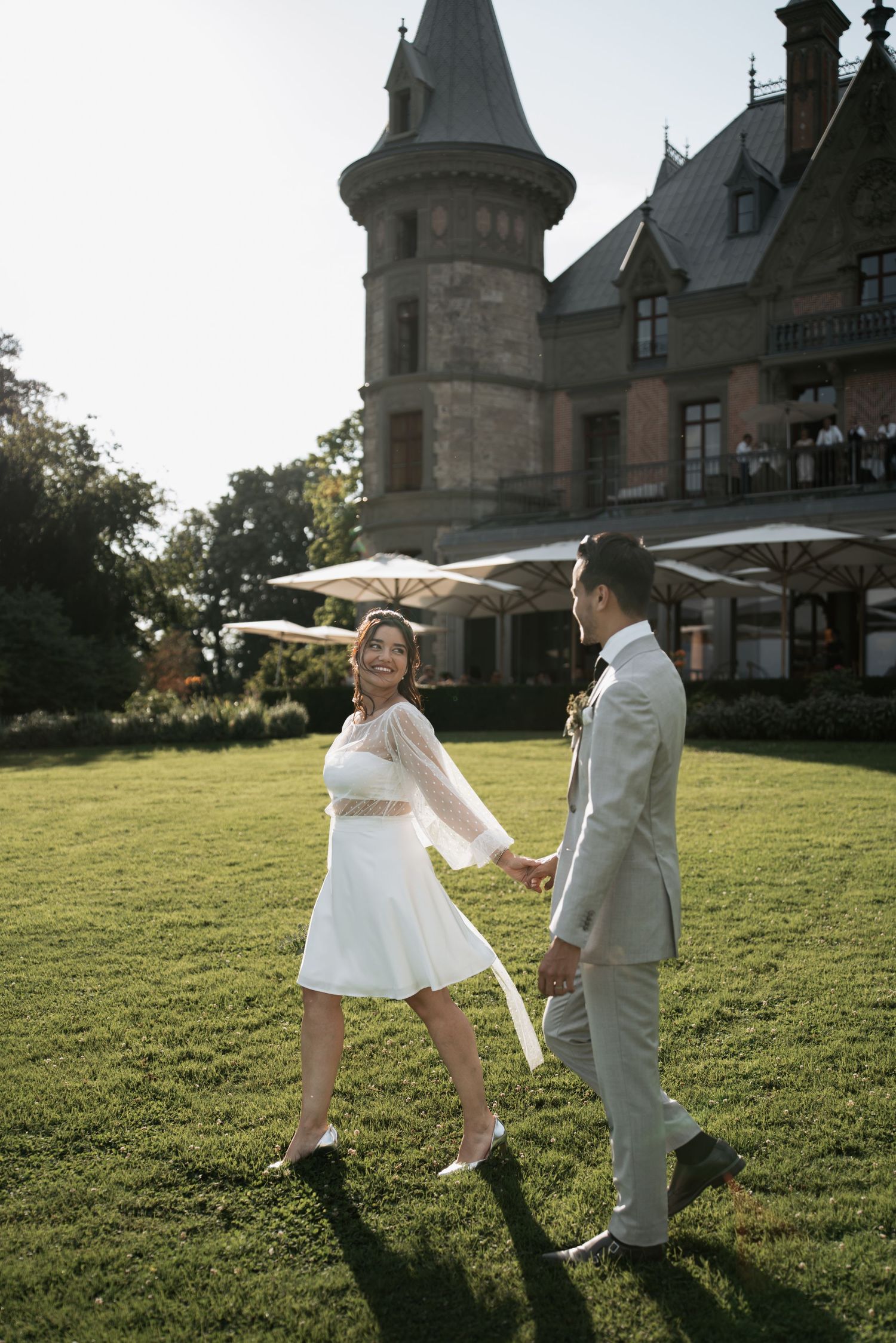 A playful wedding couple dances with parasols on a sunlit lawn near a historic castle.