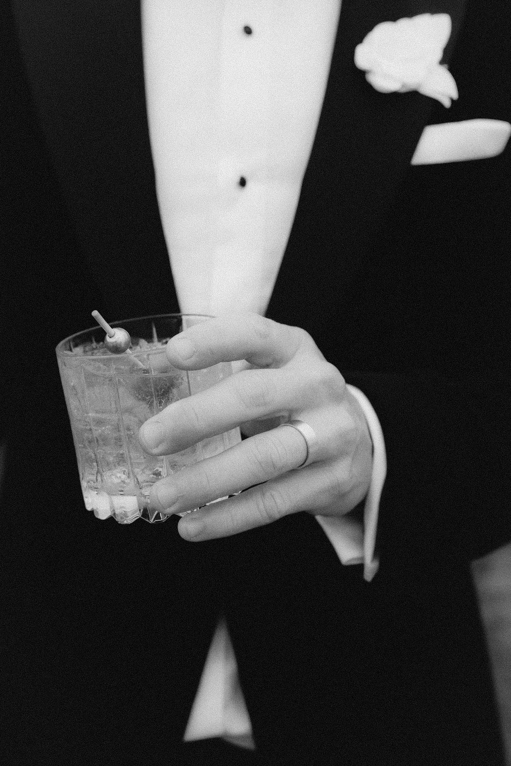 Black and white photo of someone in a tuxedo holding a cocktail with a wedding ring visible.