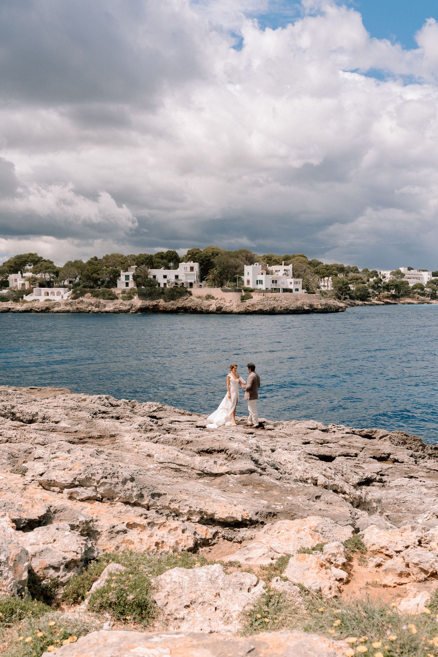 Scenic rocky coastline with white buildings in the distance and turquoise waters under cloudy skies.