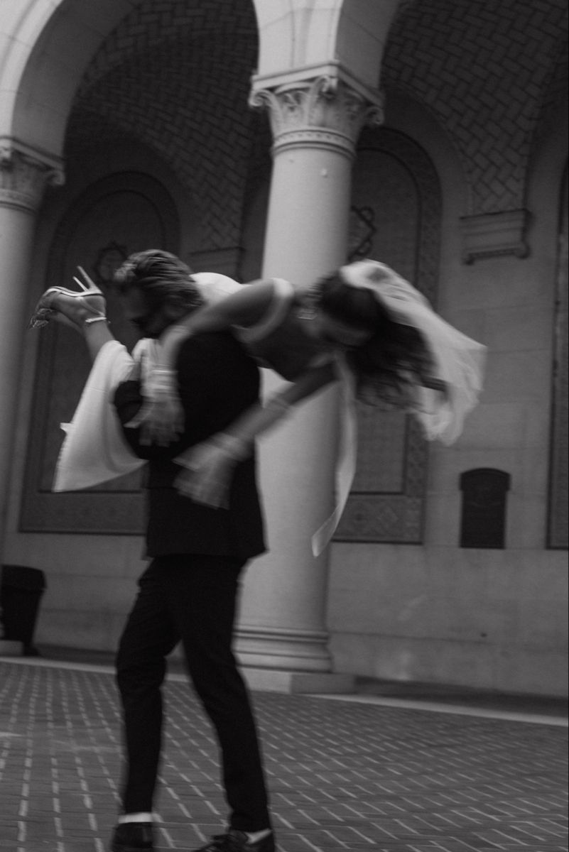 Artistic black and white shot of a dancing couple in formal wear beneath arched columns.