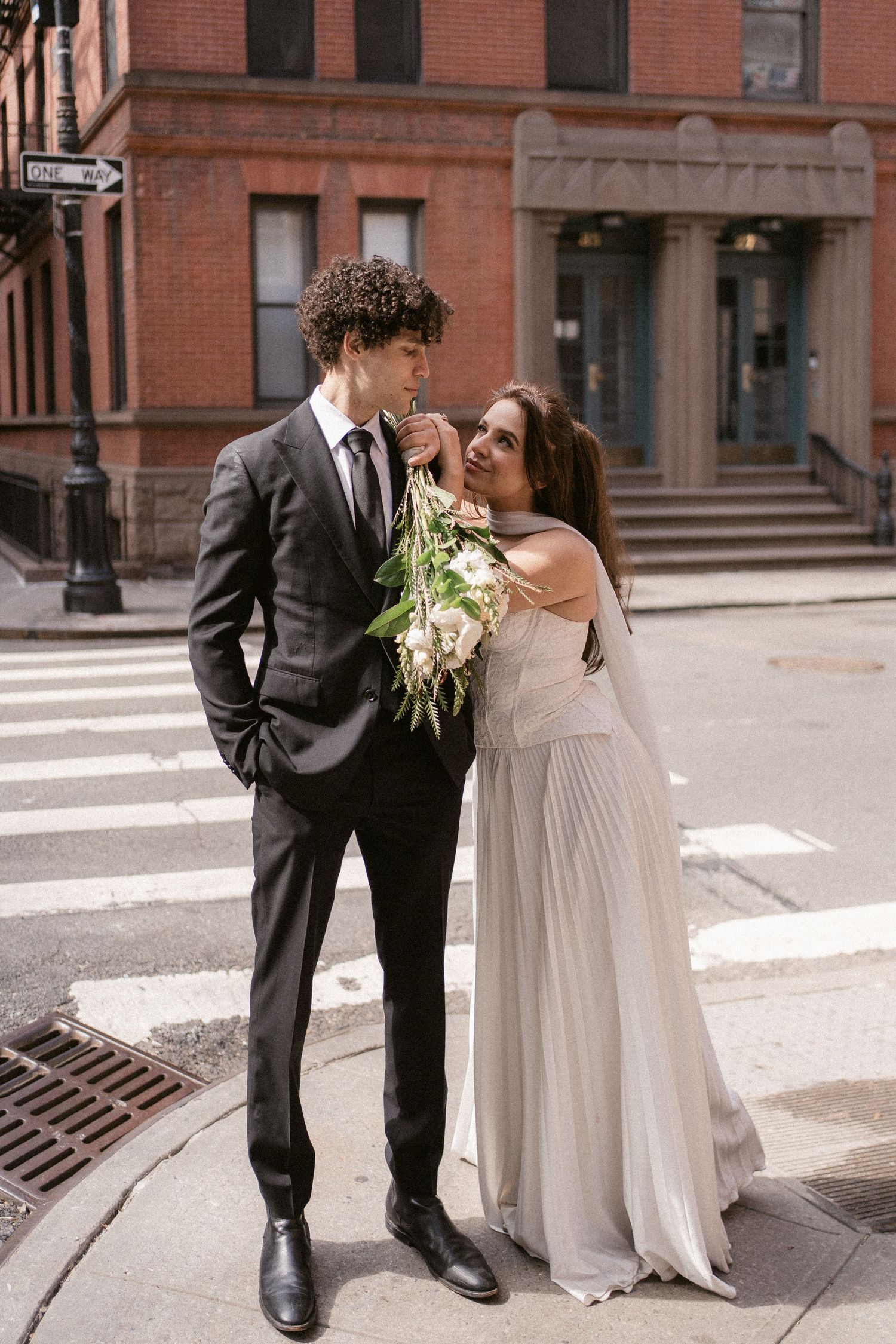 A couple in wedding attire shares an intimate moment on city streets with red brick buildings in background.