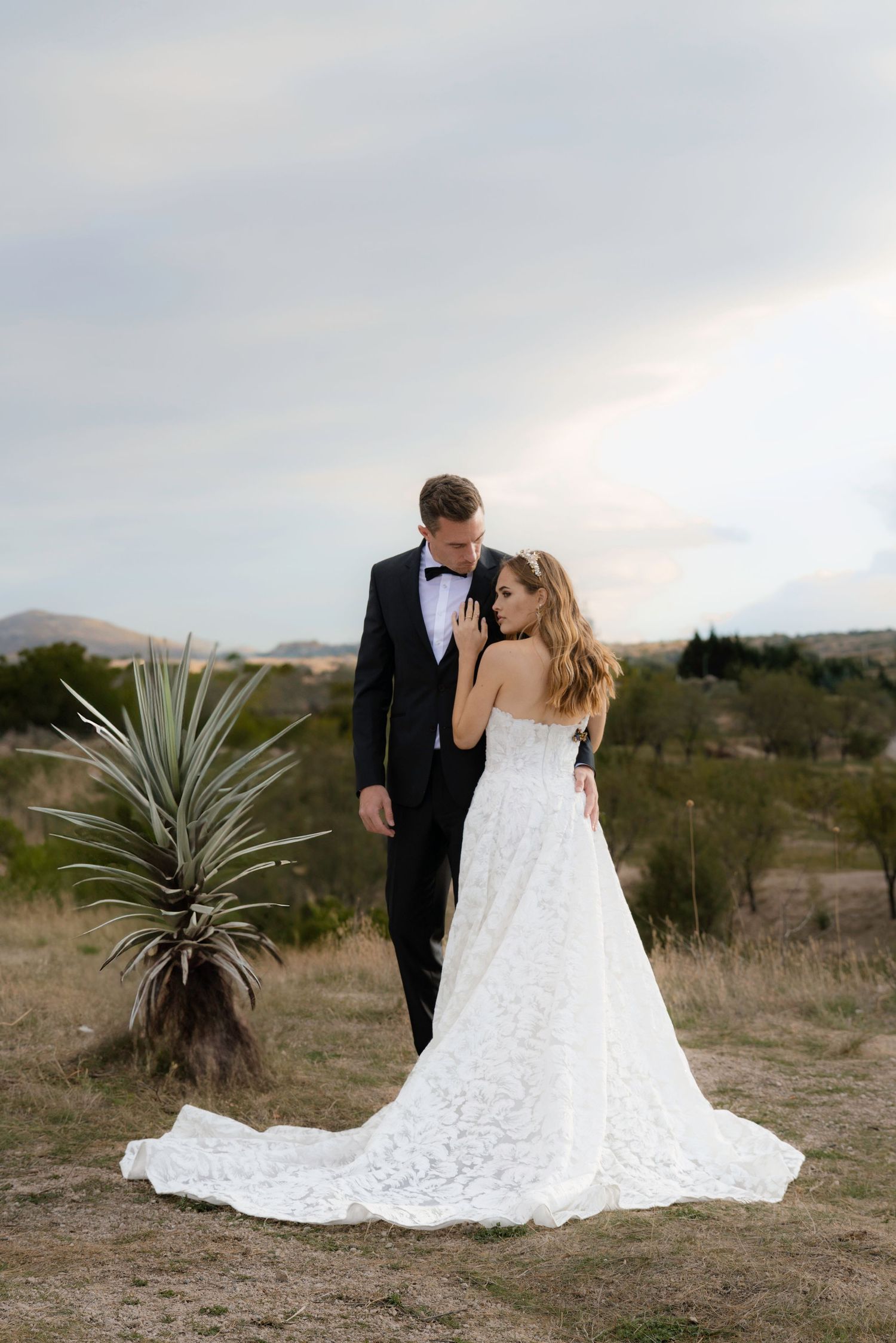 A newlywed couple stands together in a desert landscape at sunset with a white lace wedding gown.