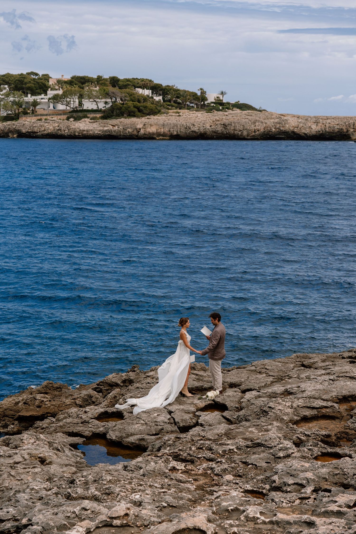 A wedding couple stands together on dramatic rocky coastline with blue waters and seaside homes in background.