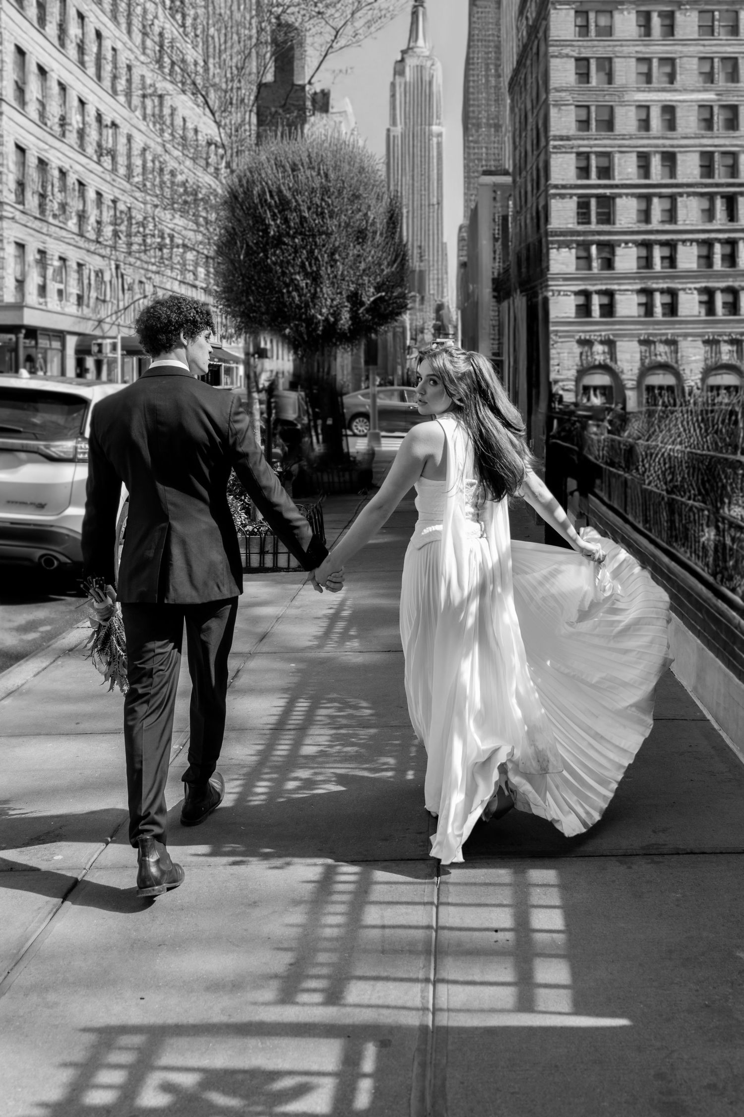A couple walking along a city sidewalk with flowing white dress and urban backdrop.