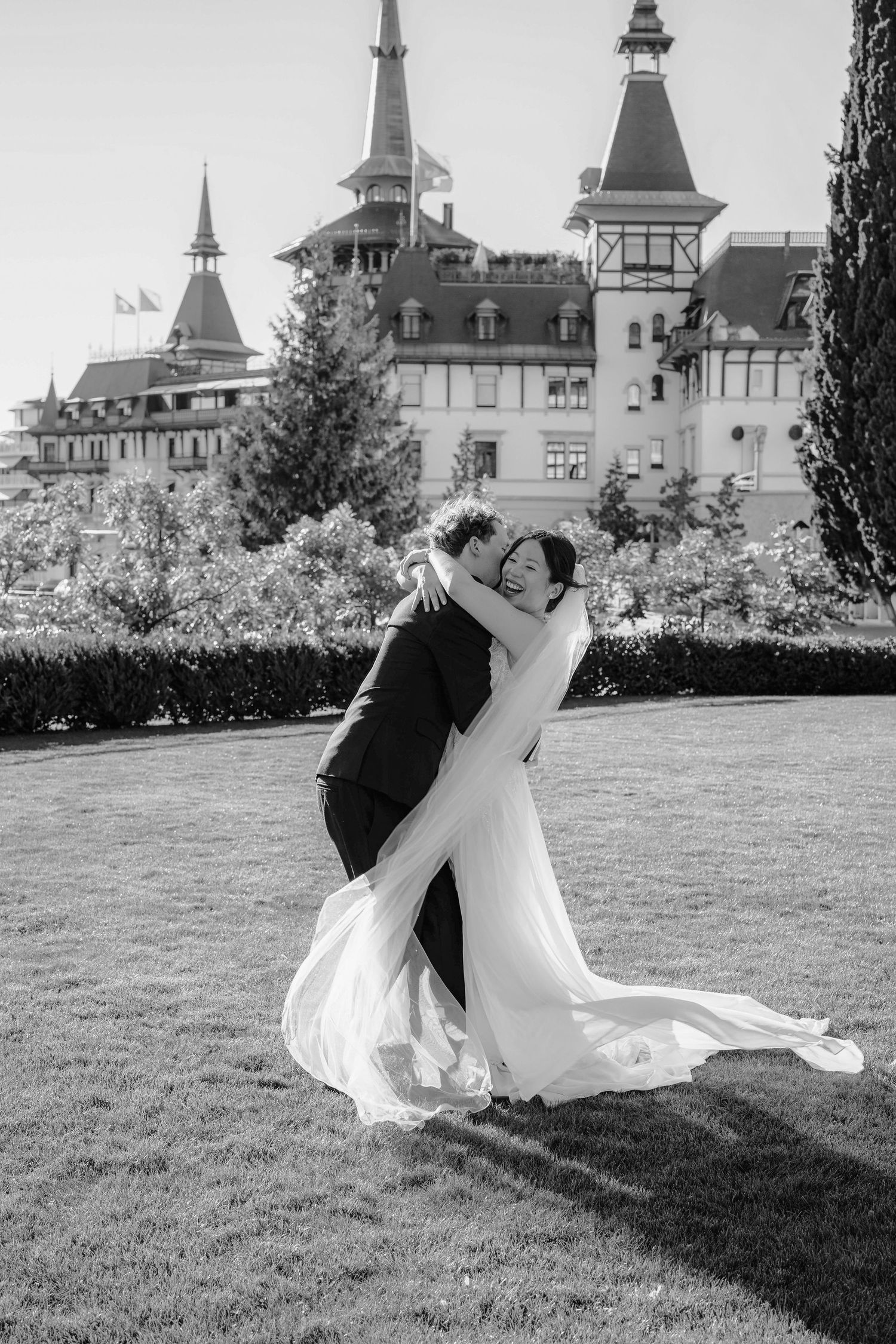 Bride in a flowing white veil embraces her groom on a manicured lawn in front of an elegant castle-style hotel in black and white.