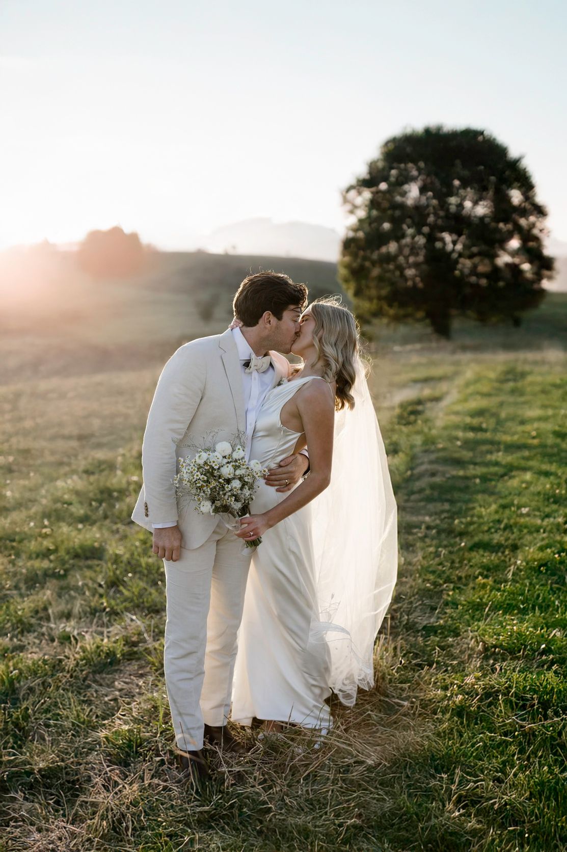 Romantic wedding couple embracing in a sunlit field with a majestic tree in the background.