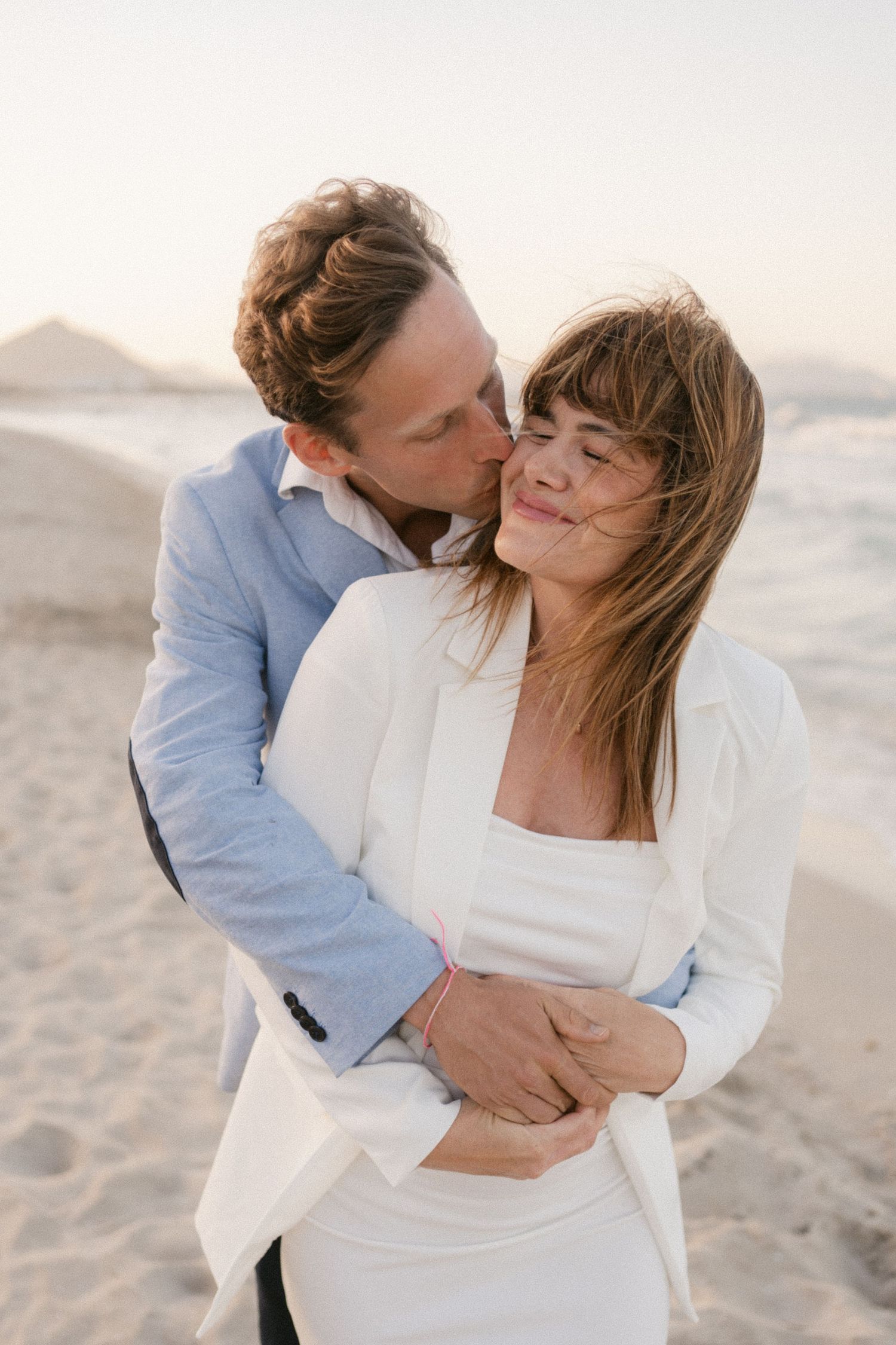 Couple in white and blue outfits sharing romantic moments on beach at sunset.