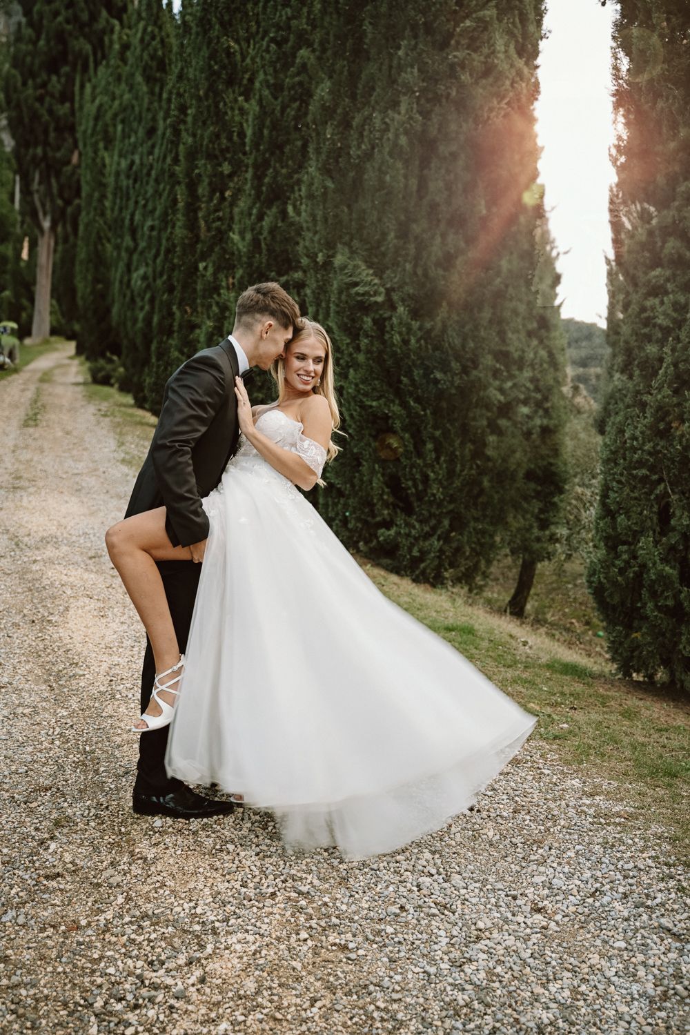 Playful wedding couple posing on gravel path surrounded by tall cypress trees.