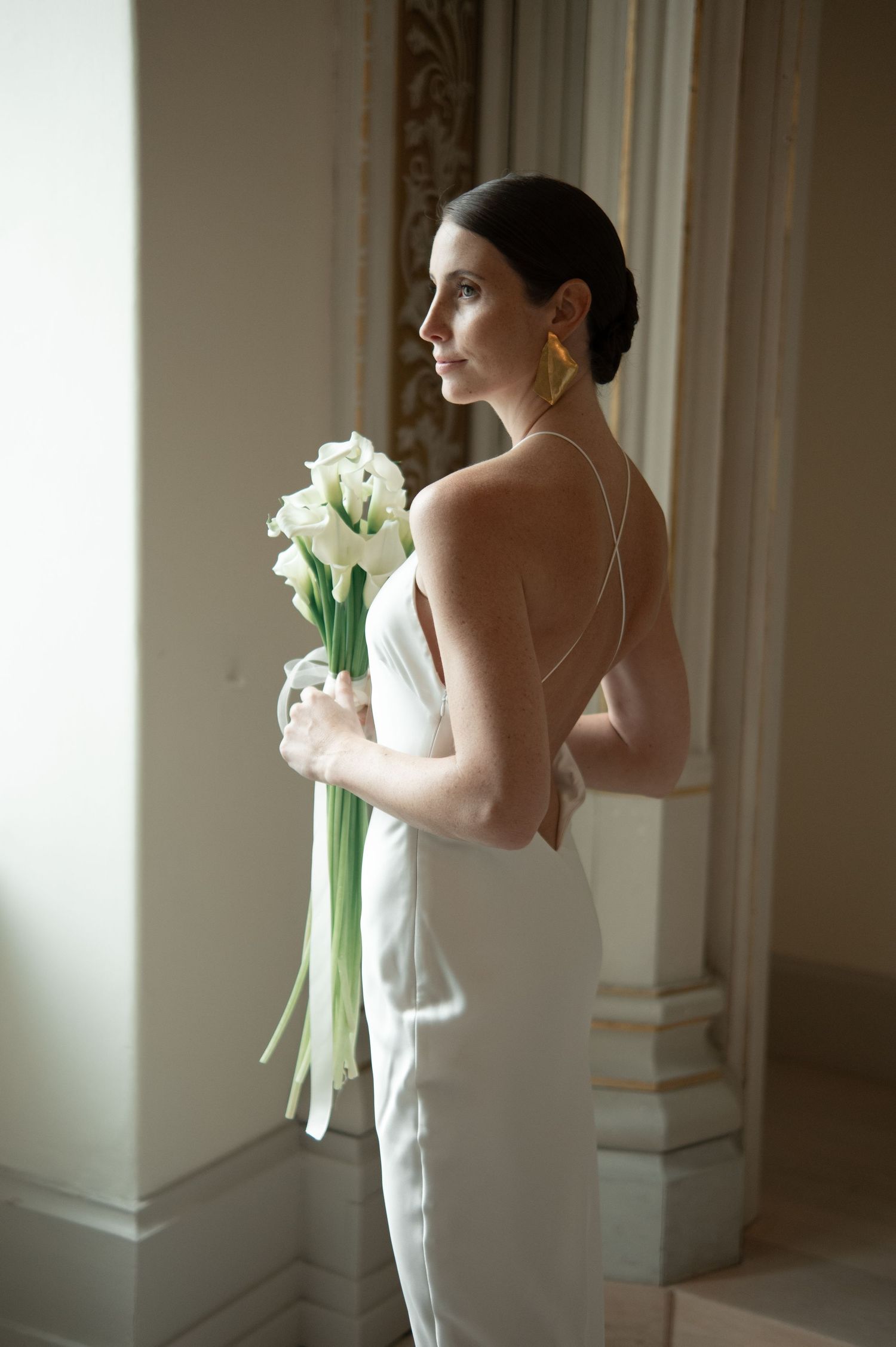 Elegant figure in white holding calla lily bouquet in soft interior lighting.