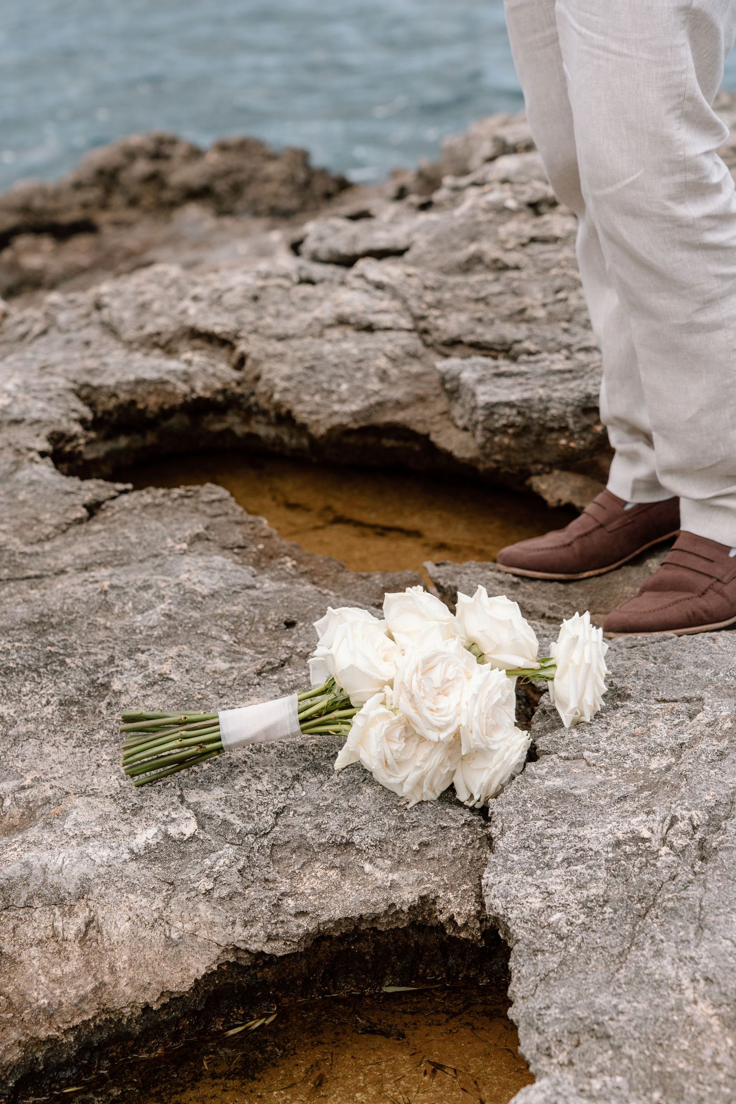 White flowers and greenery being placed near a rocky coastal edge with blue water visible.