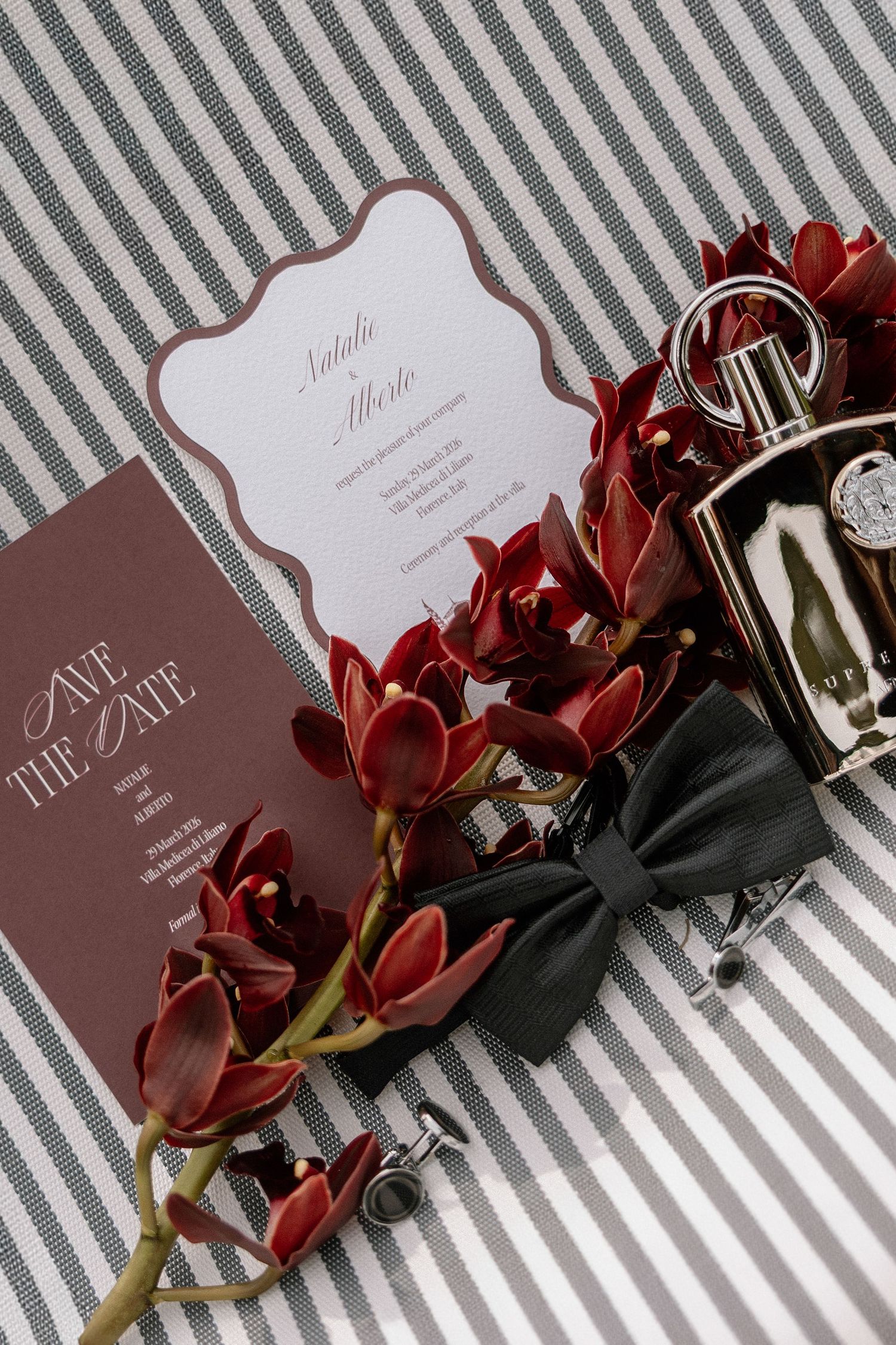 Burgundy save the date card and wedding invitation with red orchids, a black bow tie, cufflinks and perfume bottle on a striped backdrop.