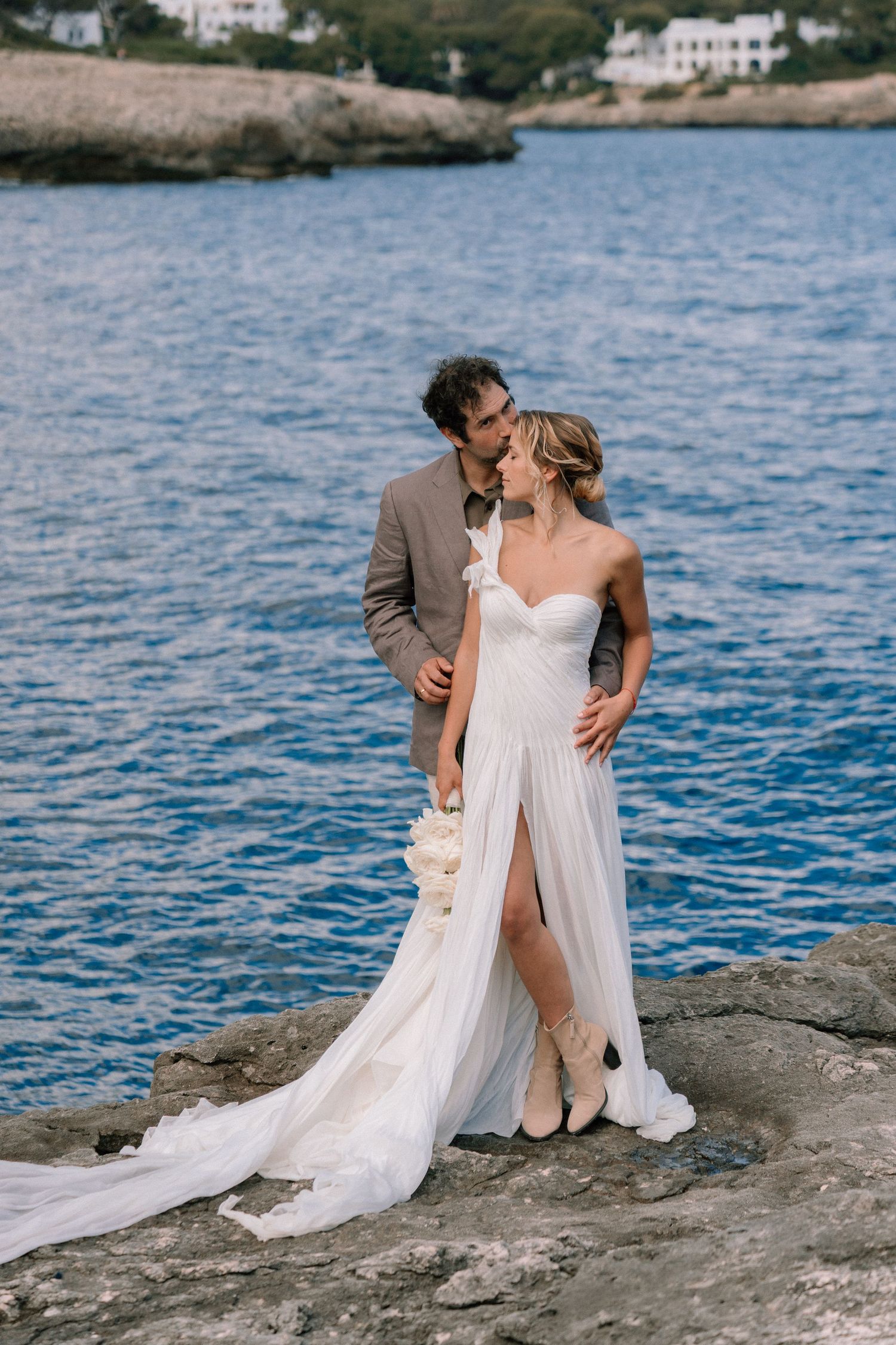 Romantic seaside wedding photoshoot on rocky coastal edge with blue water backdrop.