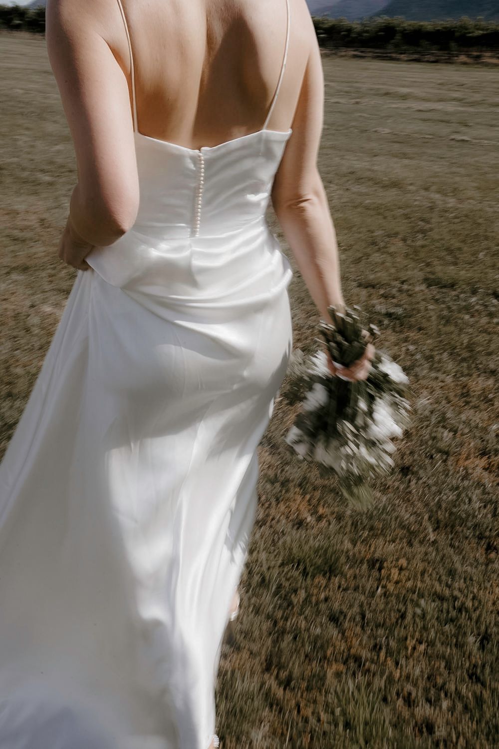 Back view of a bride in a sleek white wedding dress holding a bouquet in an outdoor setting.