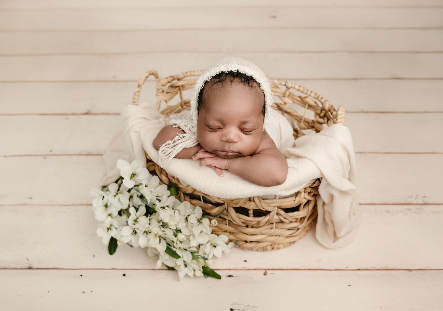 Sleeping newborn baby in a wicker basket with white flowers and cream knit bonnet on white wood background.