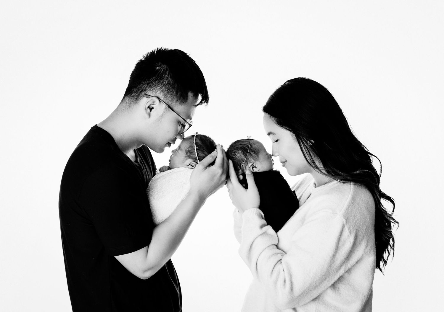 Black and white photo of couple kissing their newborn babies in a minimalist studio setting.