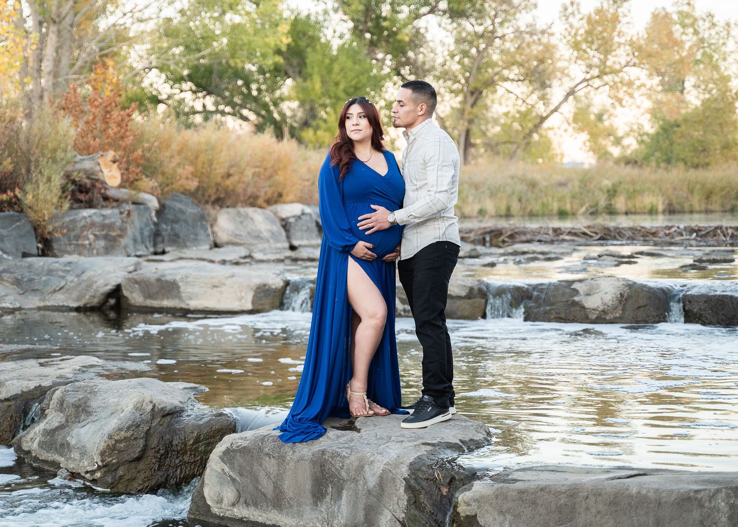 Maternity photo shoot on stone formations by water with dramatic blue dress and natural scenery.