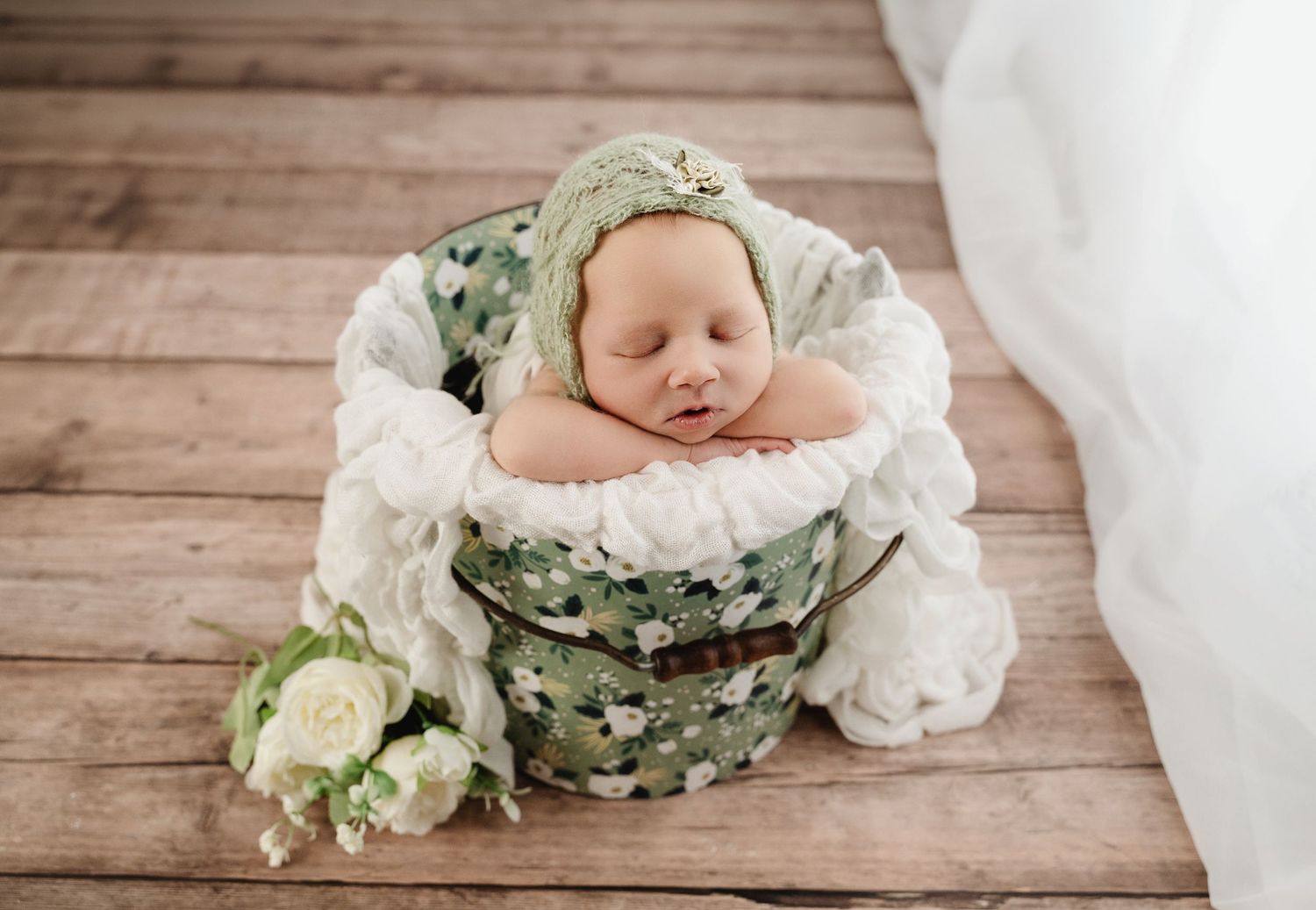A sleeping newborn nestled in a rustic wooden bowl with soft green and white floral decorations on a wooden floor.