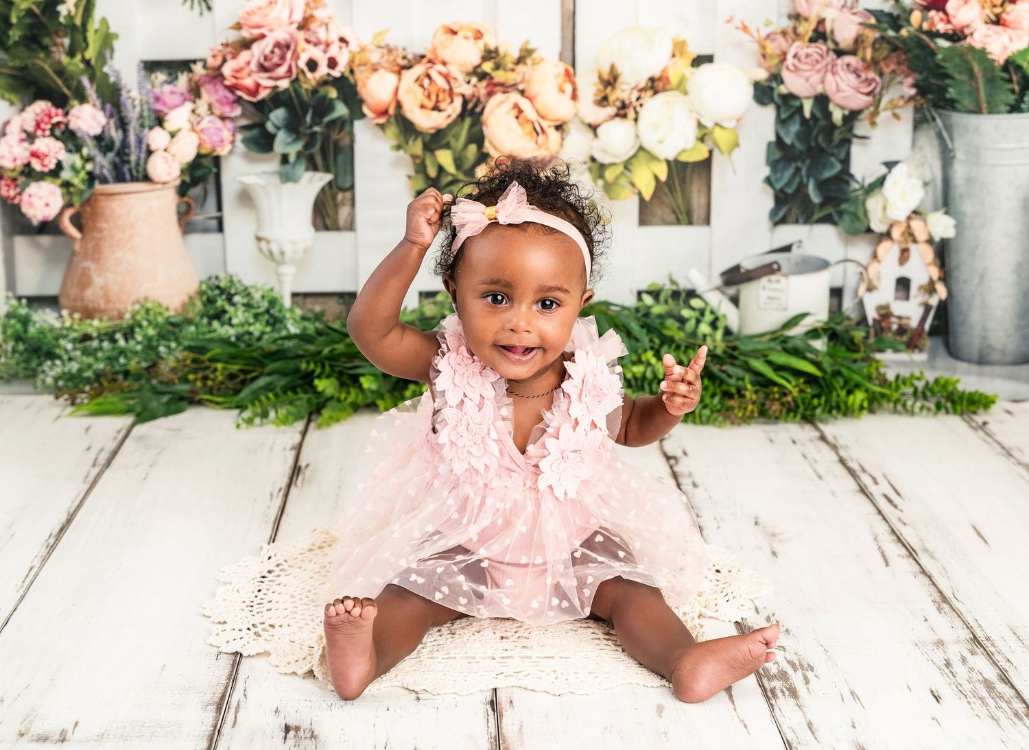 Smiling baby girl in pink lace dress sits on white wood floor with floral backdrop for first birthday photo.