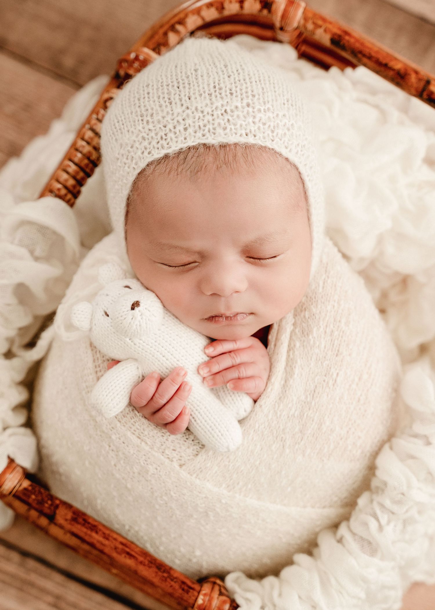A sleeping newborn wrapped in soft white knit blankets rests peacefully in a rustic wooden basket.