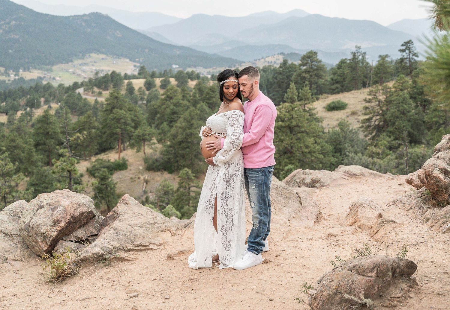 A couple sits together on a rocky mountain overlook with scenic mountain views in the background.