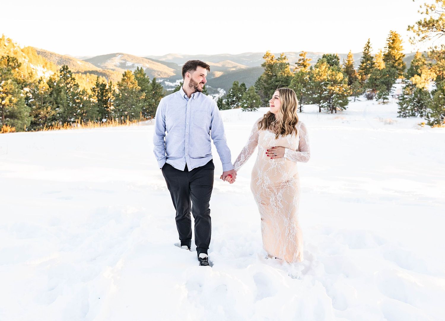 A couple walks hand in hand through a snowy mountain landscape at sunset with golden light filtering through trees.