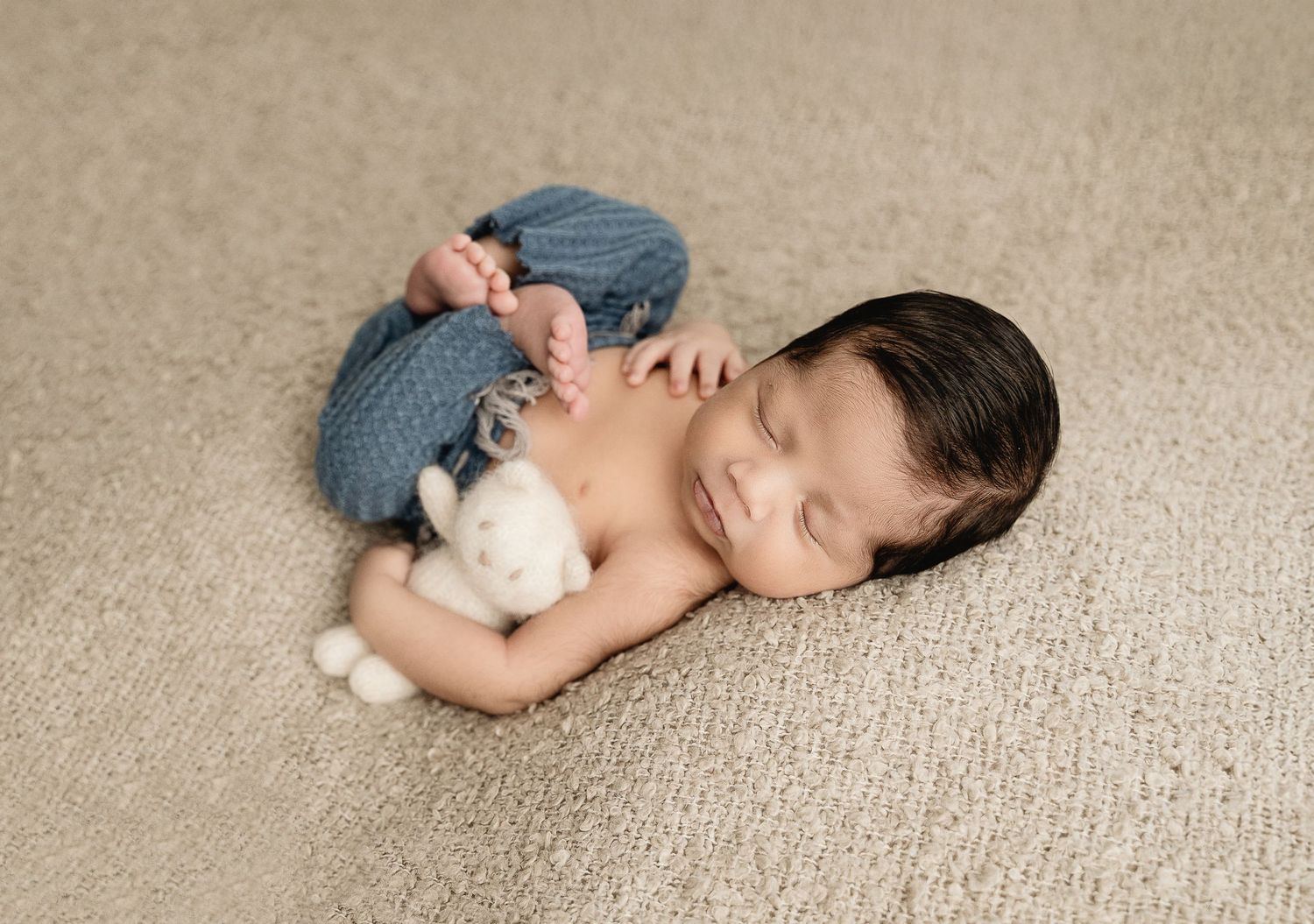 A peaceful sleeping newborn wearing blue pants and curled up on a beige textured carpet.