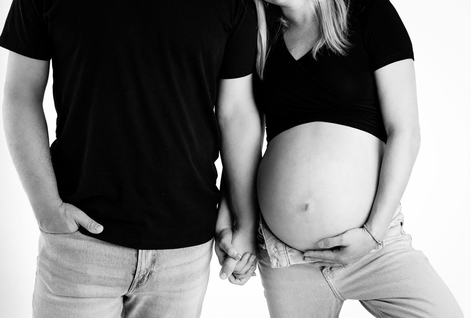Black and white maternity photo of pregnant couple holding hands, showing baby bump, wearing casual jeans and black tops.
