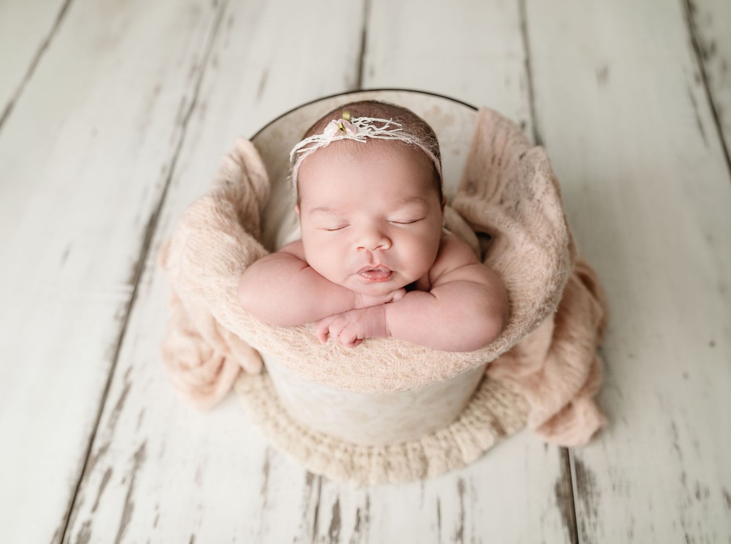 Newborn baby girl sleeping peacefully in a bucket with soft pink blanket and delicate tiara headband.