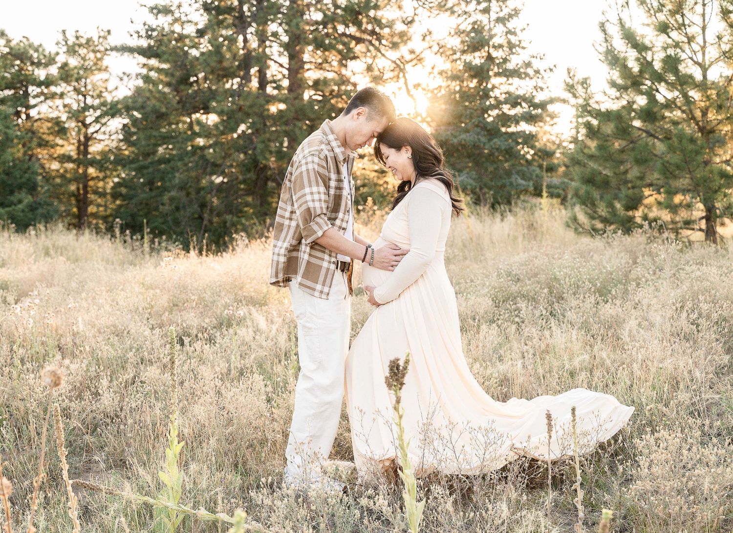 A romantic couple embraces in a sunlit meadow with tall grass and evergreen trees at golden hour.