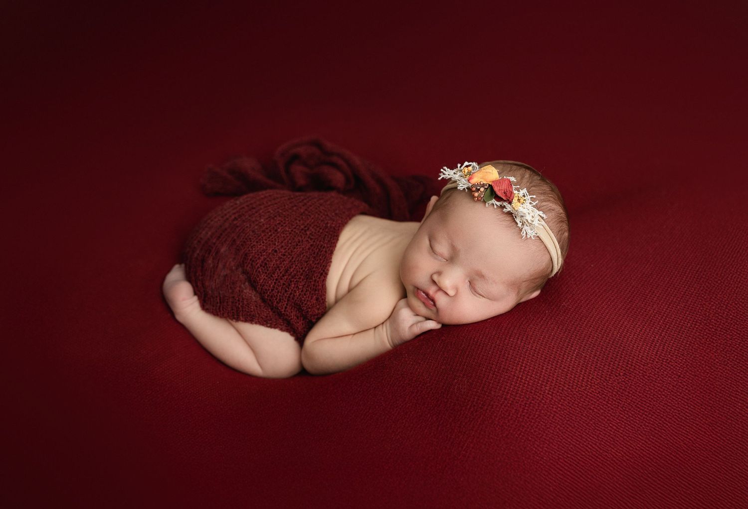 Sleeping newborn wearing burgundy wrap and floral headband poses on matching red backdrop for professional portrait.