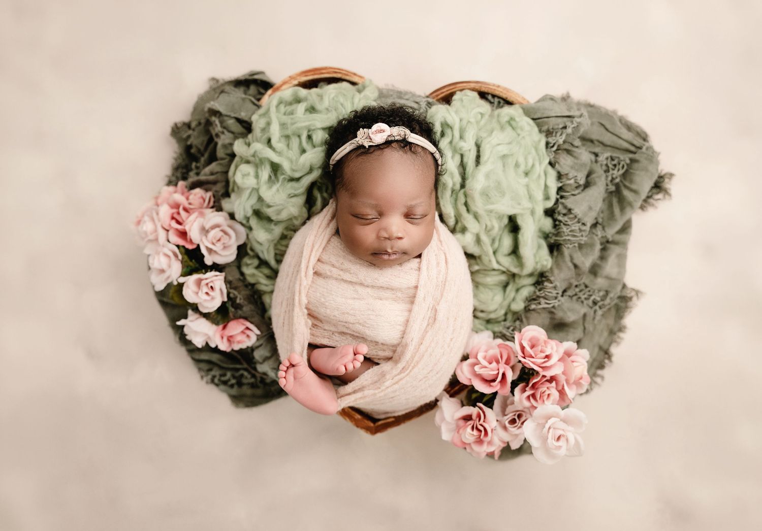 Newborn baby sleeping in a wooden bowl with pink roses and green fabric for a professional photography session.