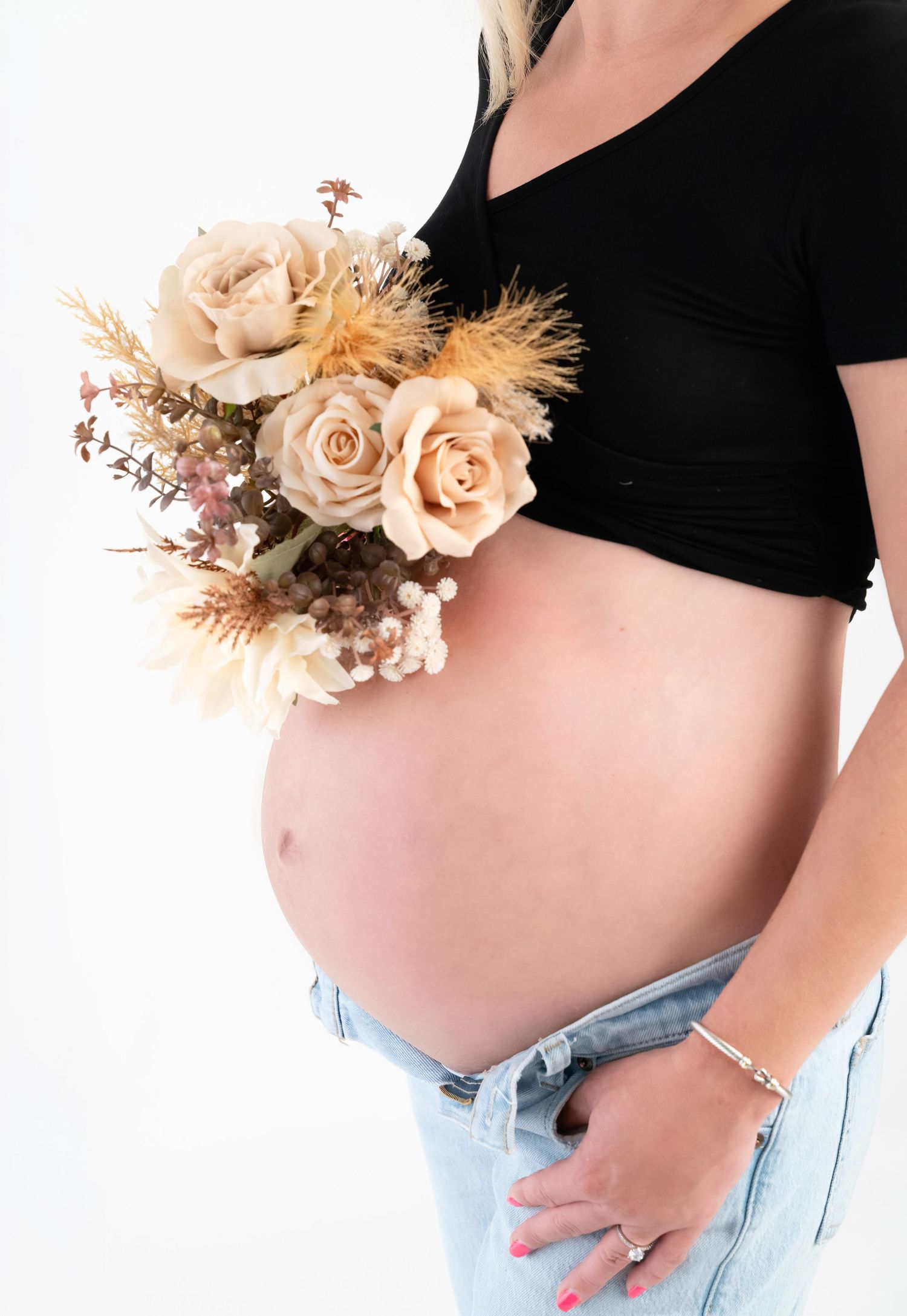 Pregnant woman in black crop top and jeans holding a bouquet of beige roses, showcasing her baby bump.