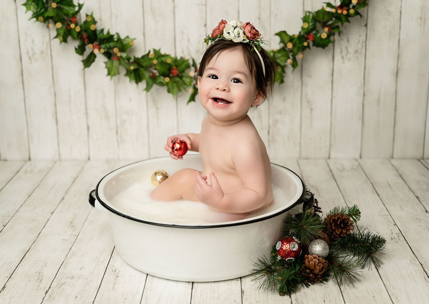 Holiday themed baby portrait in white enamel bath basin with evergreen garland and ornament decorations.