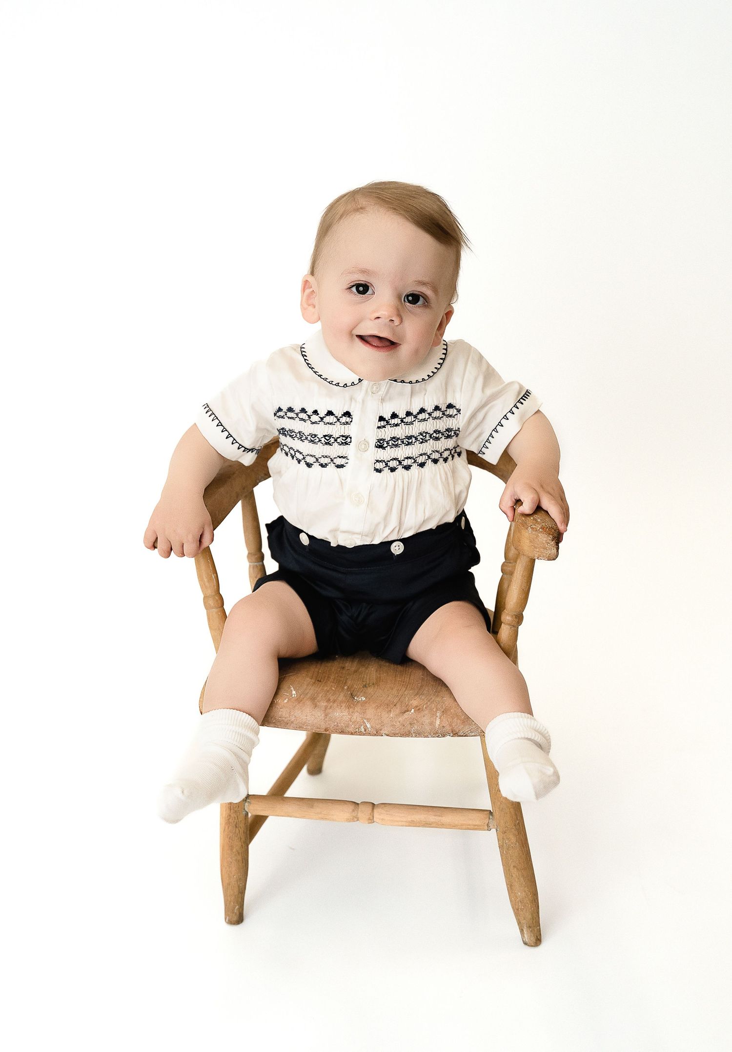 Baby in black and white outfit sits on small wooden chair during studio photoshoot.