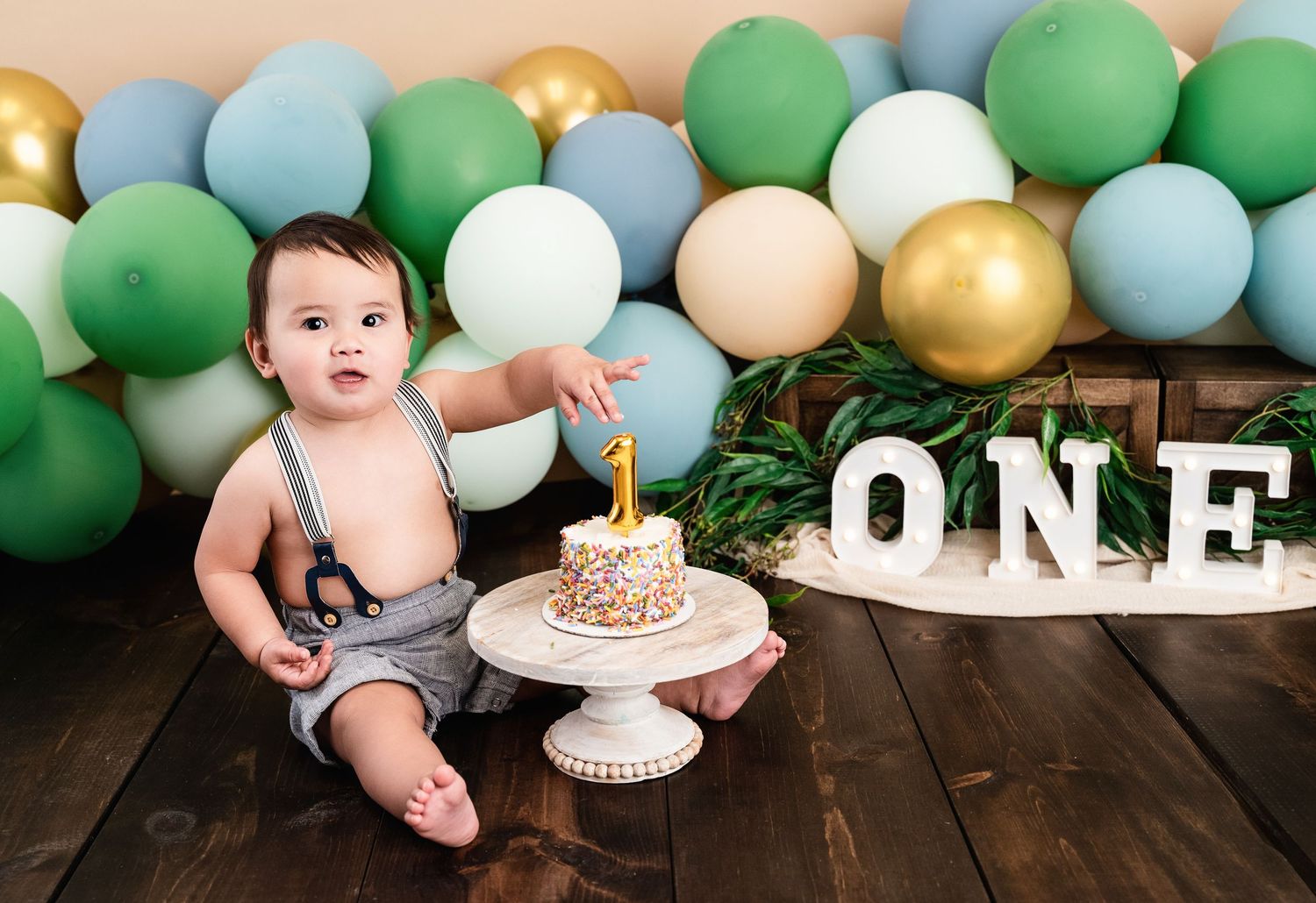 Baby boy in suspenders sits beside a smash cake at his first birthday party with green and gold balloons.